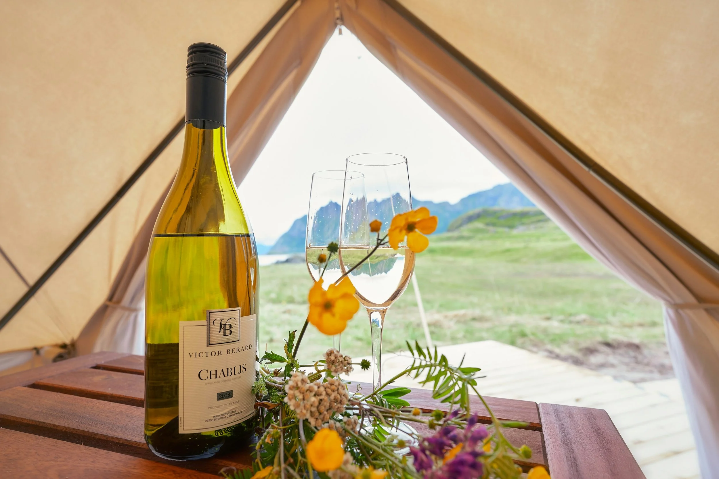 Bottle of Chablis wine and two glasses displayed inside a tent overlooking scenic countryside, representing the fresh and elegant Chardonnay wines of Chablis, France.