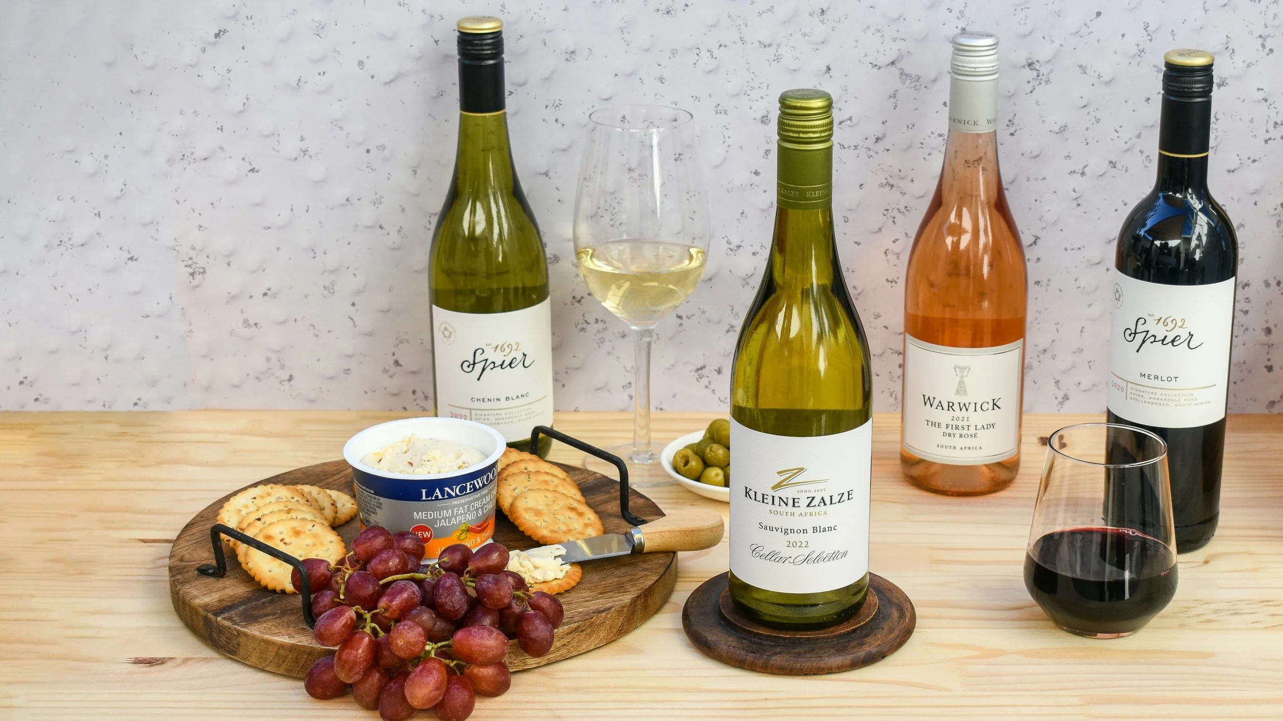 Wine bottles, glasses, crackers, cheese, and grapes arranged for a home wine tasting with food pairings.