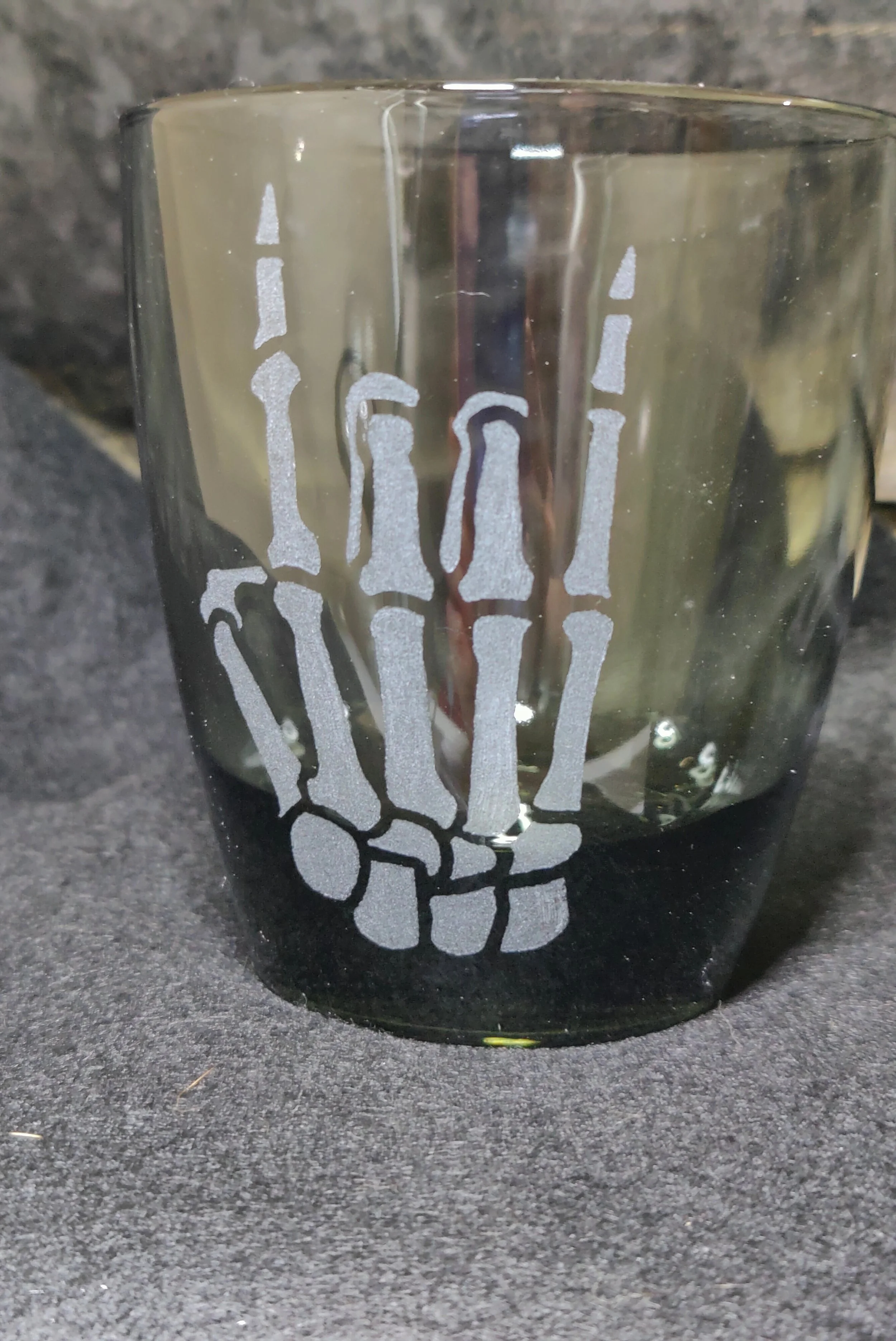 A glass with a skeleton hand design engraved on it, placed on a dark surface with a stone background.