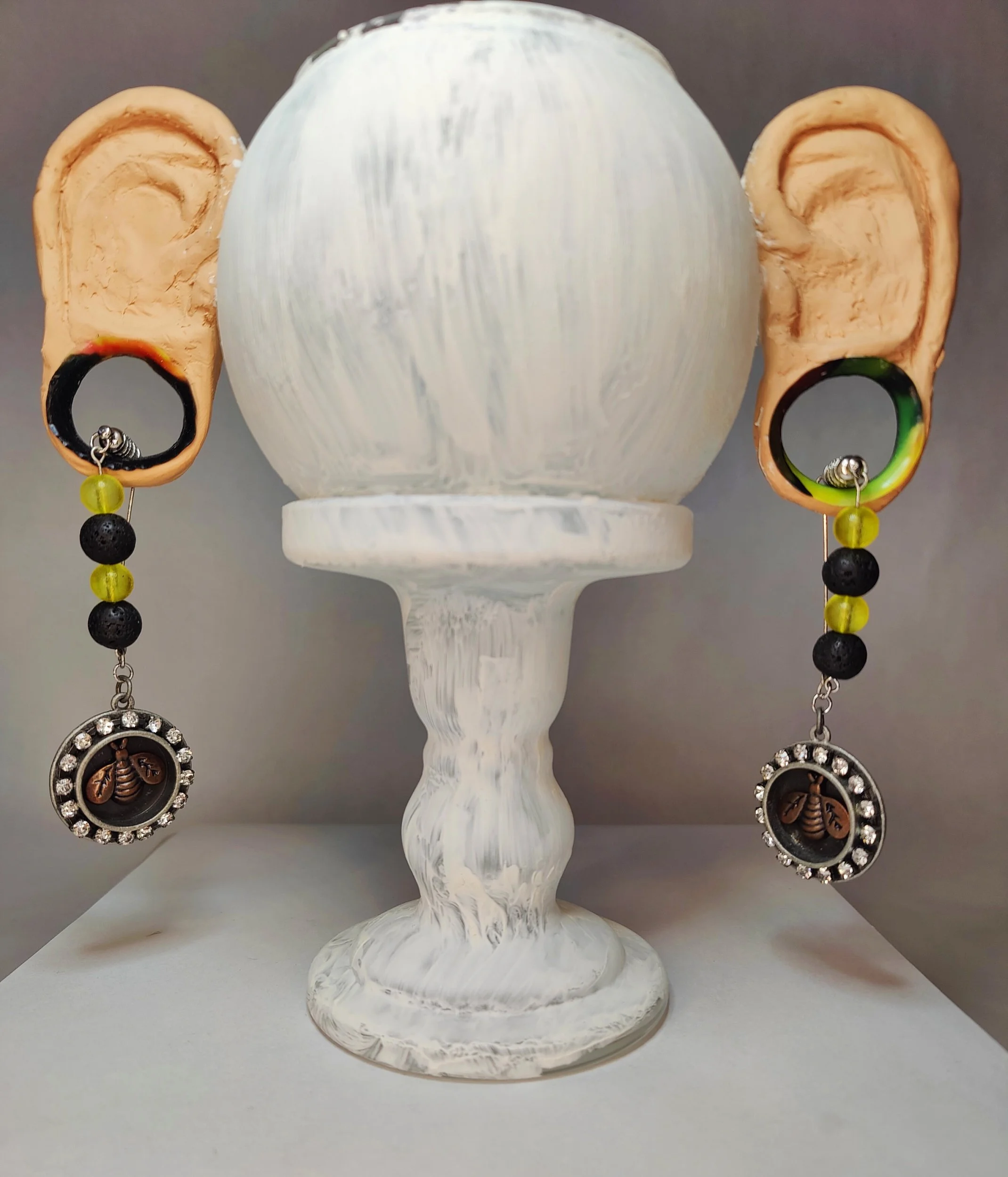 A glass display painted white with clay ears.  The ear lobes are stretched and have handmade earrings through the tunnels.
