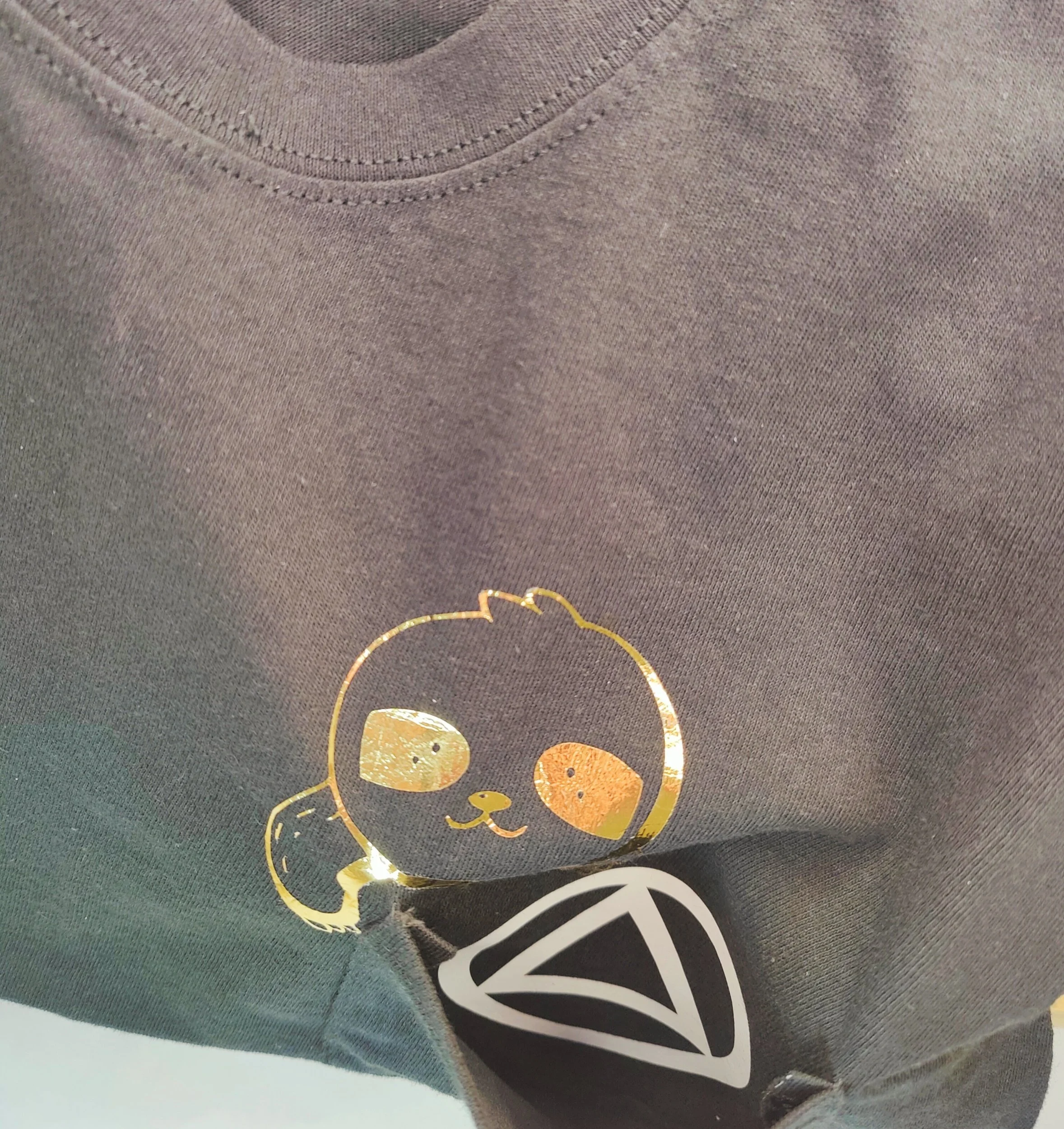 Close-up of a black T-shirt with a gold foil sloth illustration and a white recovery symbol inside a pocket.