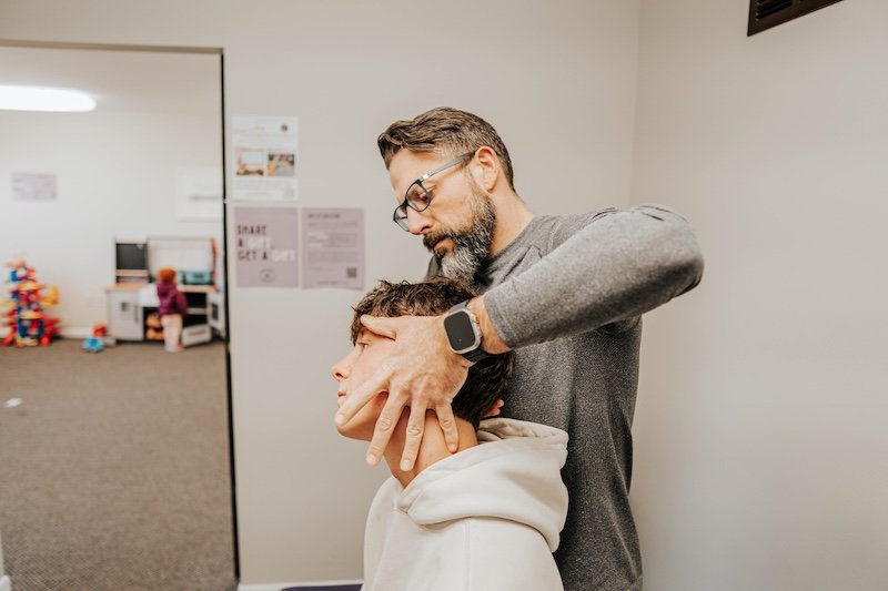 Chiropractor Dr. Jeremy Lawson adjusting a person's neck in an office setting, with another room visible where a child is playing.