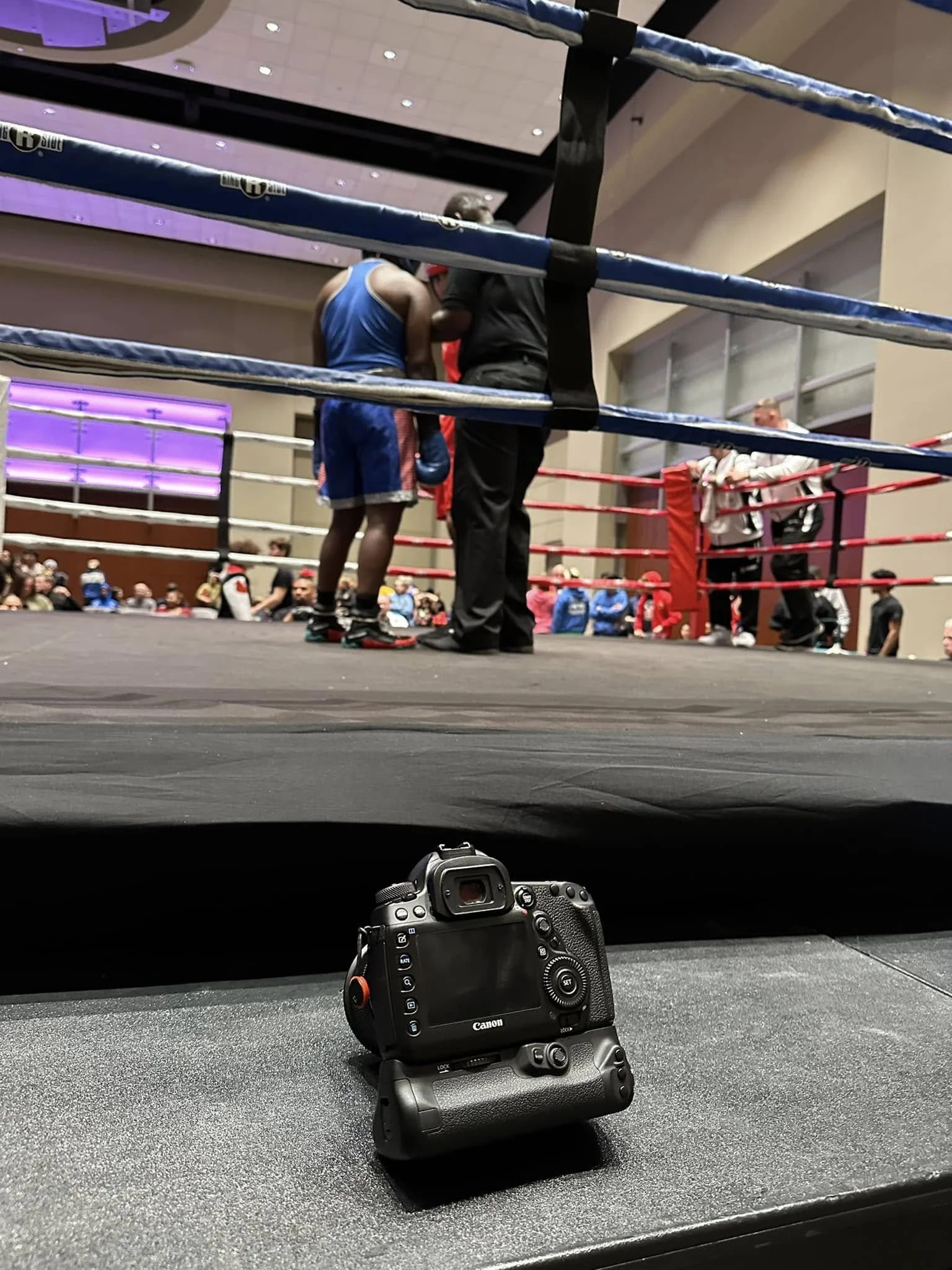 Boxing ring with two boxers and a referee, Canon camera in foreground.