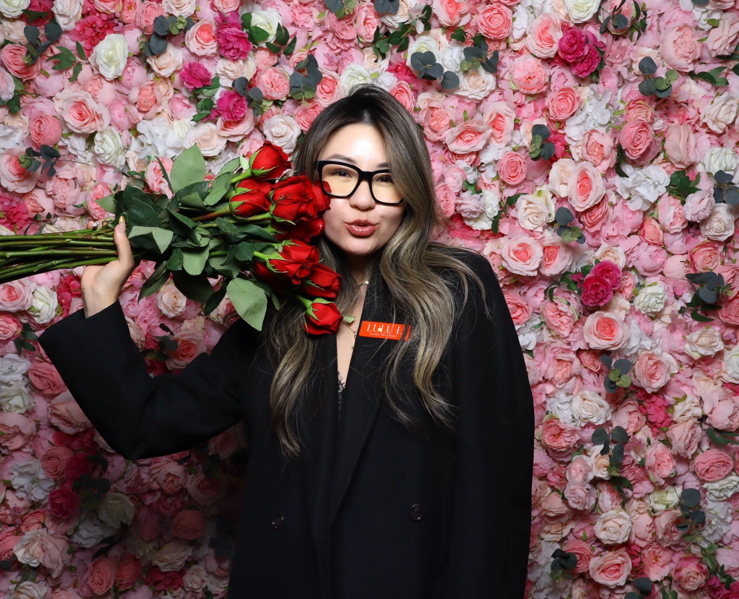 A woman with shoulder-length hair, wearing glasses and a black knitted sweater, posing with one arm over her head in front of a wall covered with pink and white roses and green leaves.