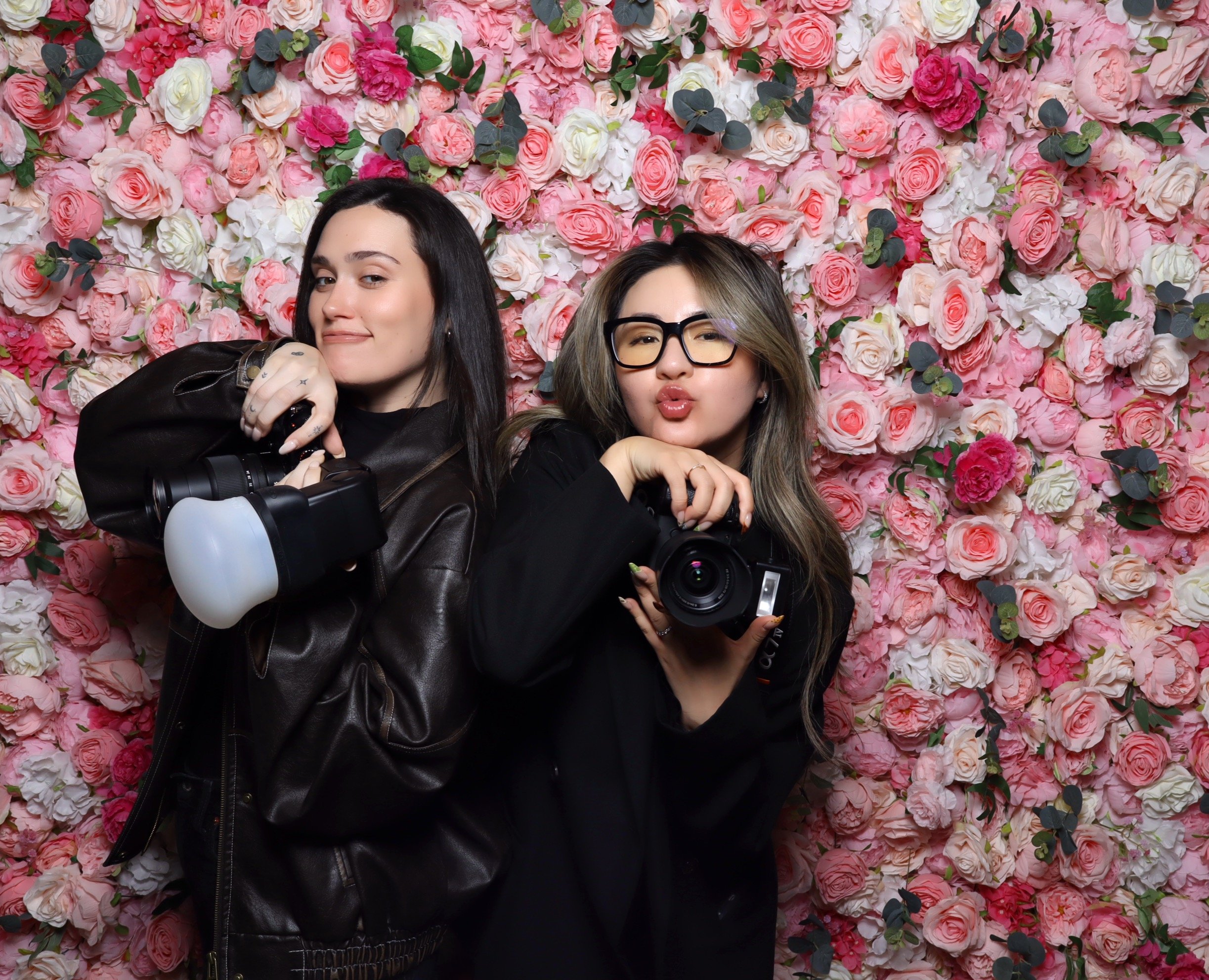 A woman with shoulder-length hair, wearing glasses and a black knitted sweater, posing with one arm over her head in front of a wall covered with pink and white roses and green leaves.