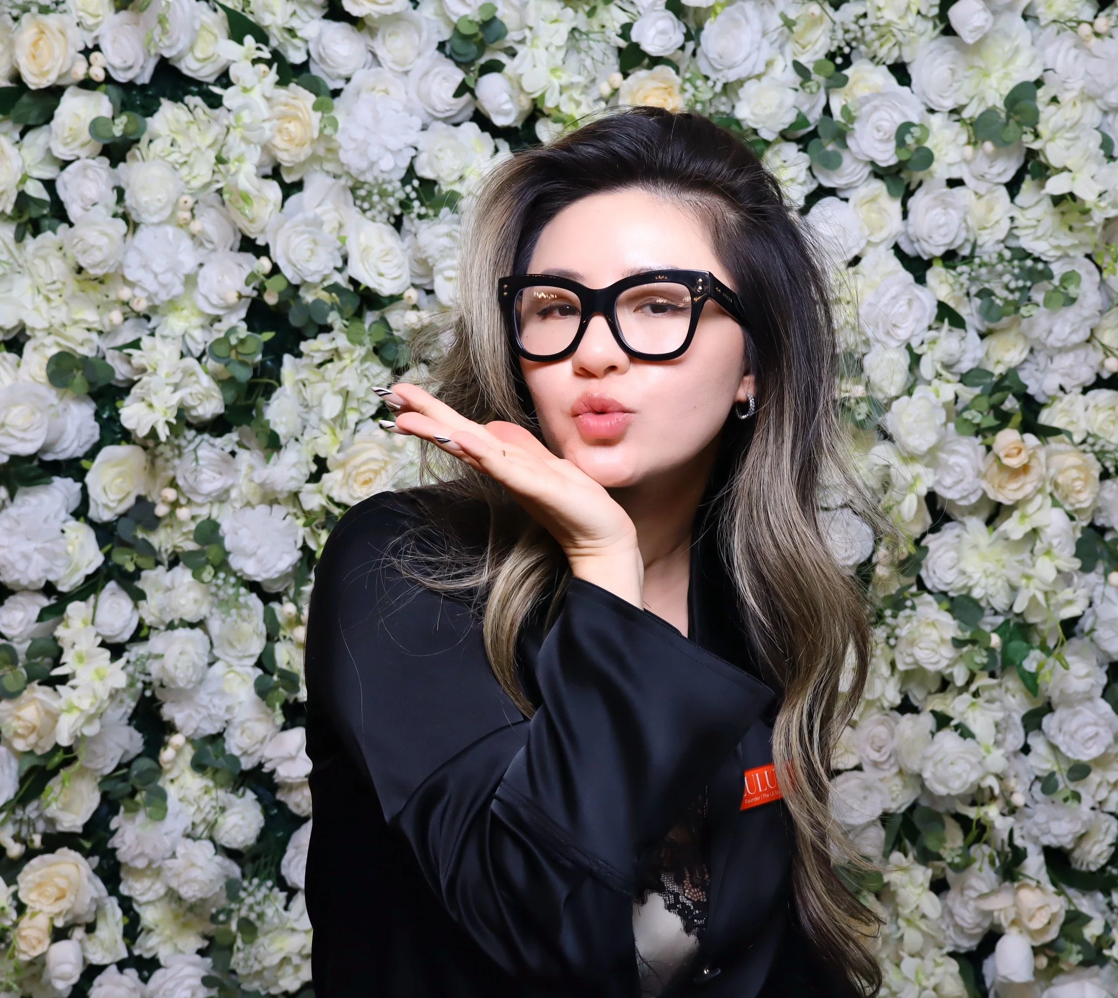 Woman with long hair and black glasses making a heart shape with her hands in front of a white floral background.