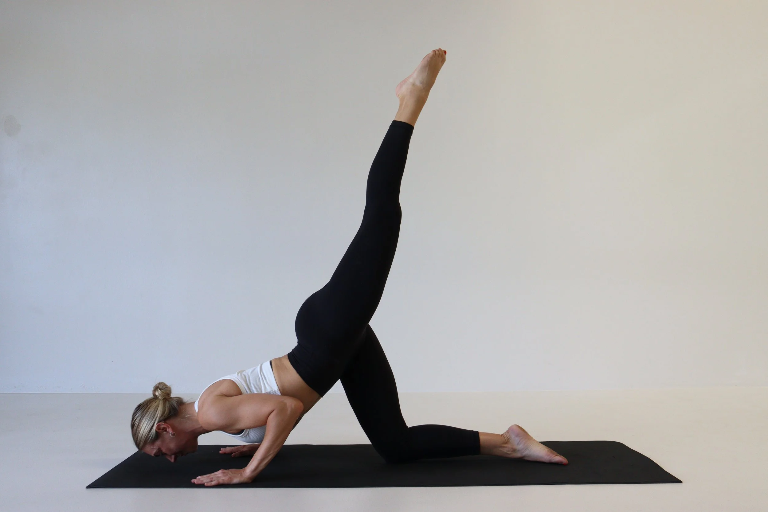 A woman practicing yoga on a black mat in a room with a plain white wall, performing a pose with one knee and one hand on the ground, one arm extended upward, and one leg raised straight back.