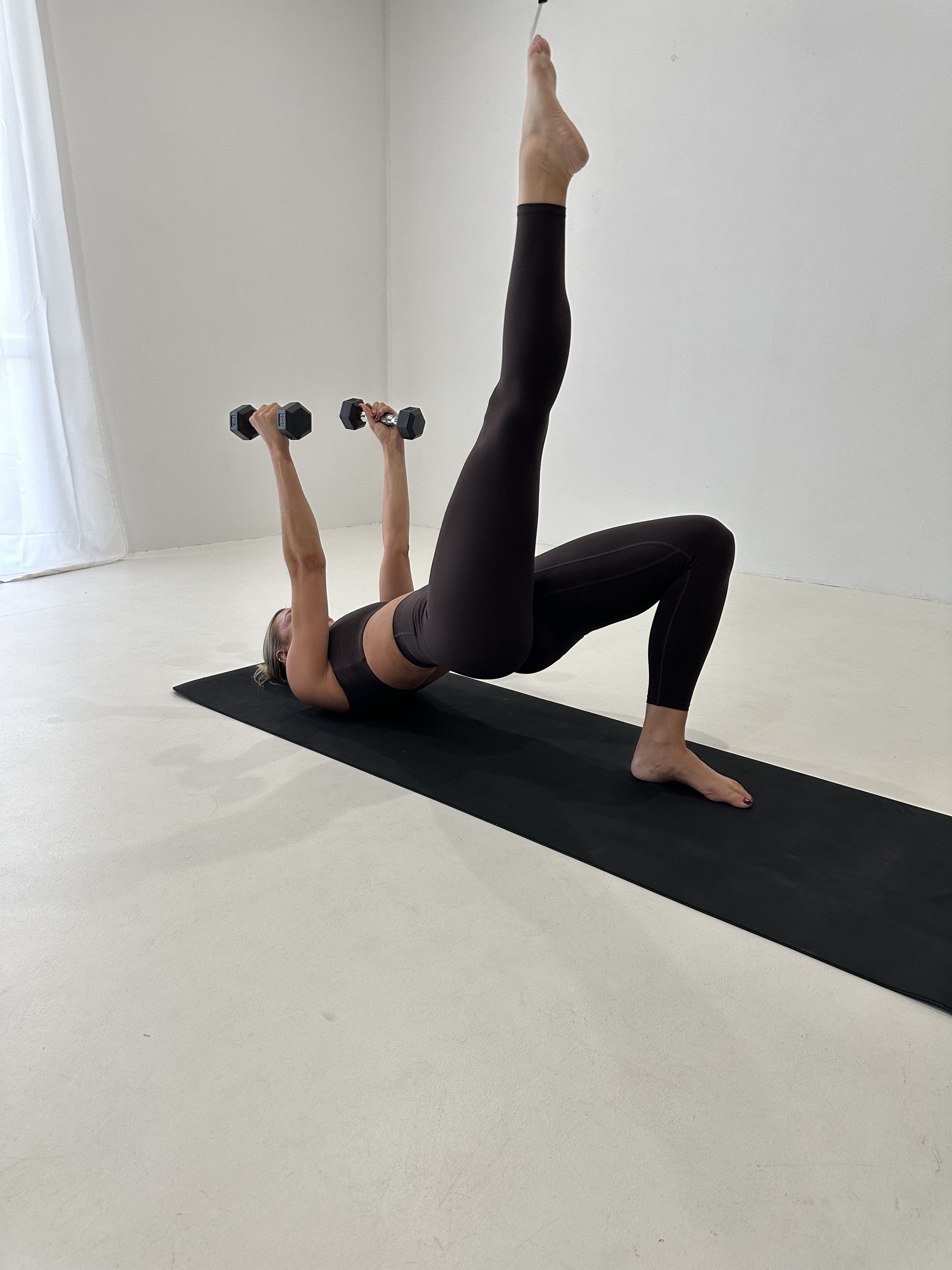 Woman lying on the floor on a black exercise mat, holding dumbbells with both hands, one leg extended straight up and the other bent, performing a workout in a bright, minimalistic room.