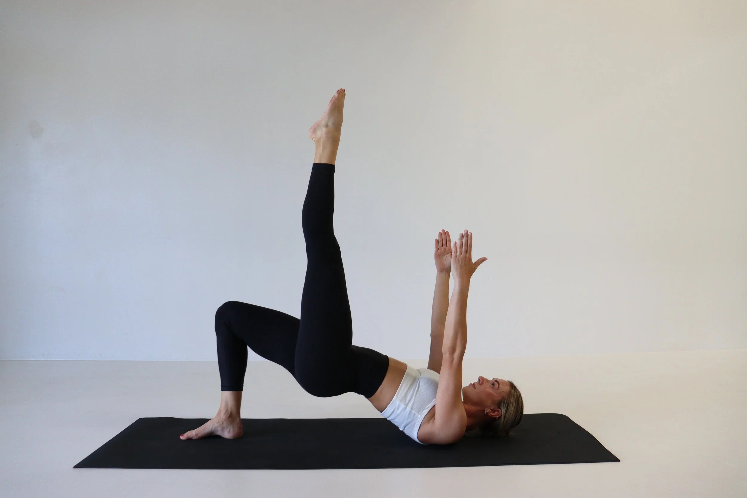 A woman practicing yoga on a black mat, lying on her back with one leg raised and arms extended upward.