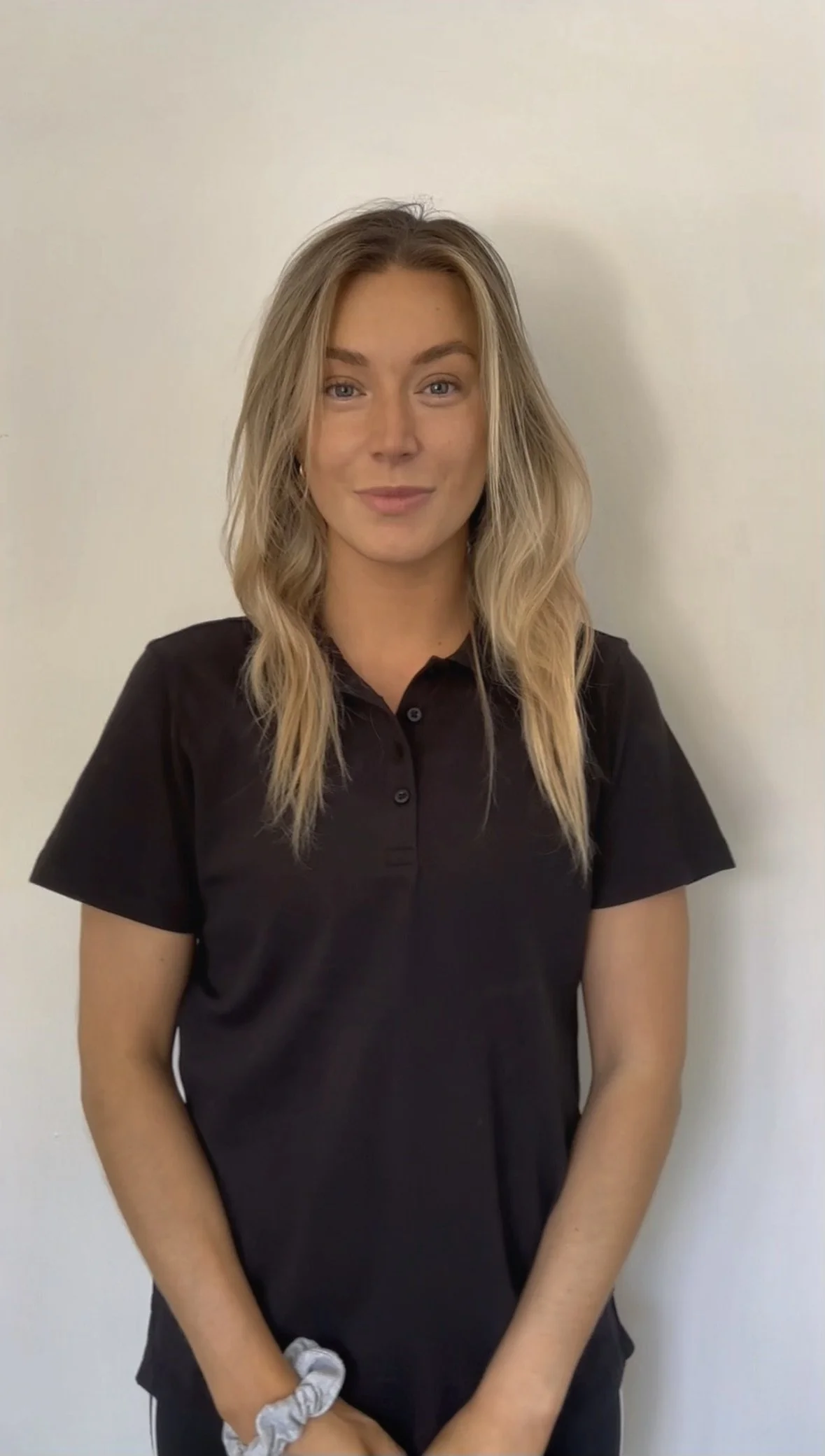 A young woman with blonde hair and blue eyes wearing a black collared shirt standing against a plain white wall.