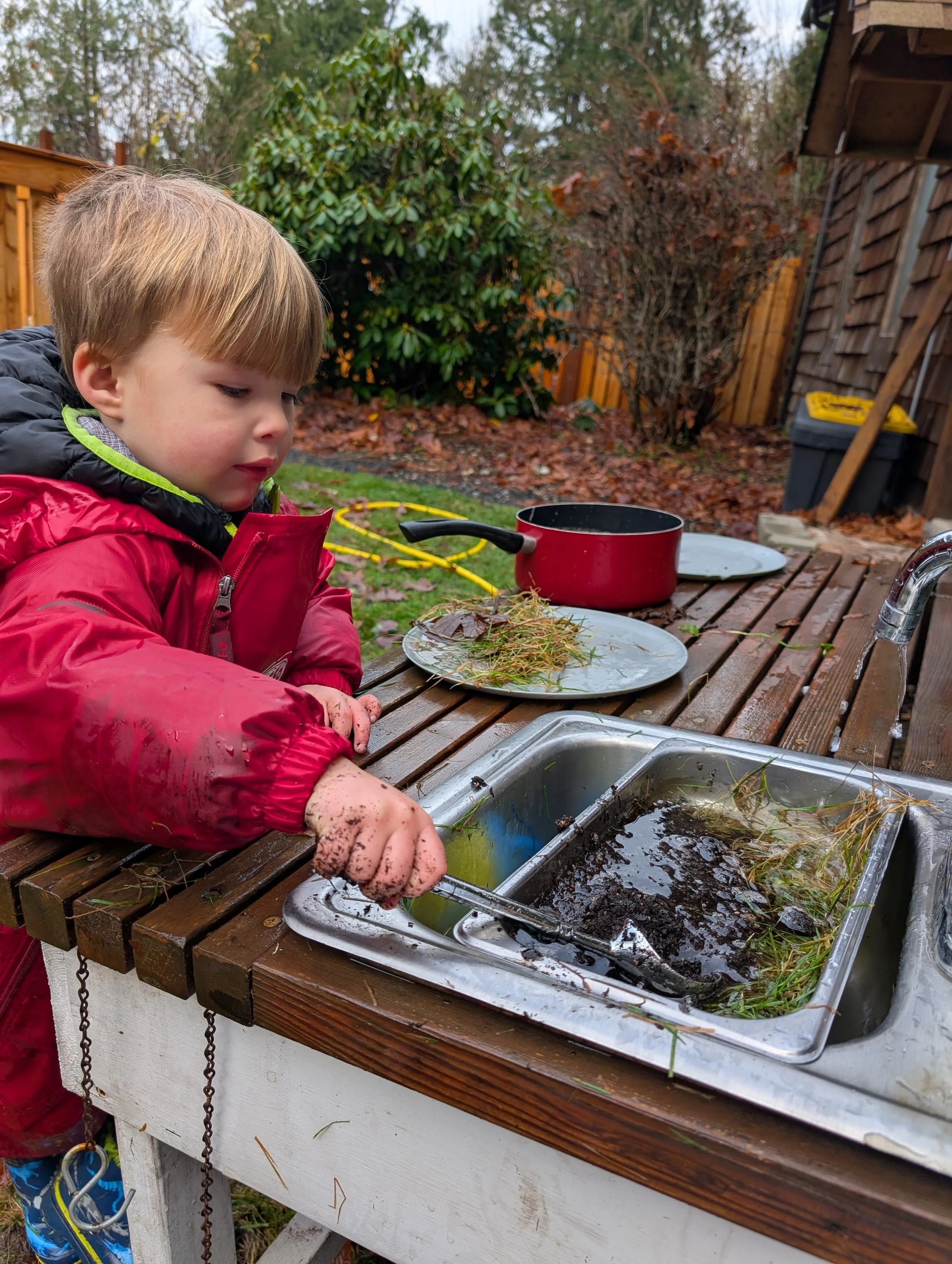 A young boy in a red jacket washing mud and plant debris in a kitchen sink outside table, with a backyard in the background.