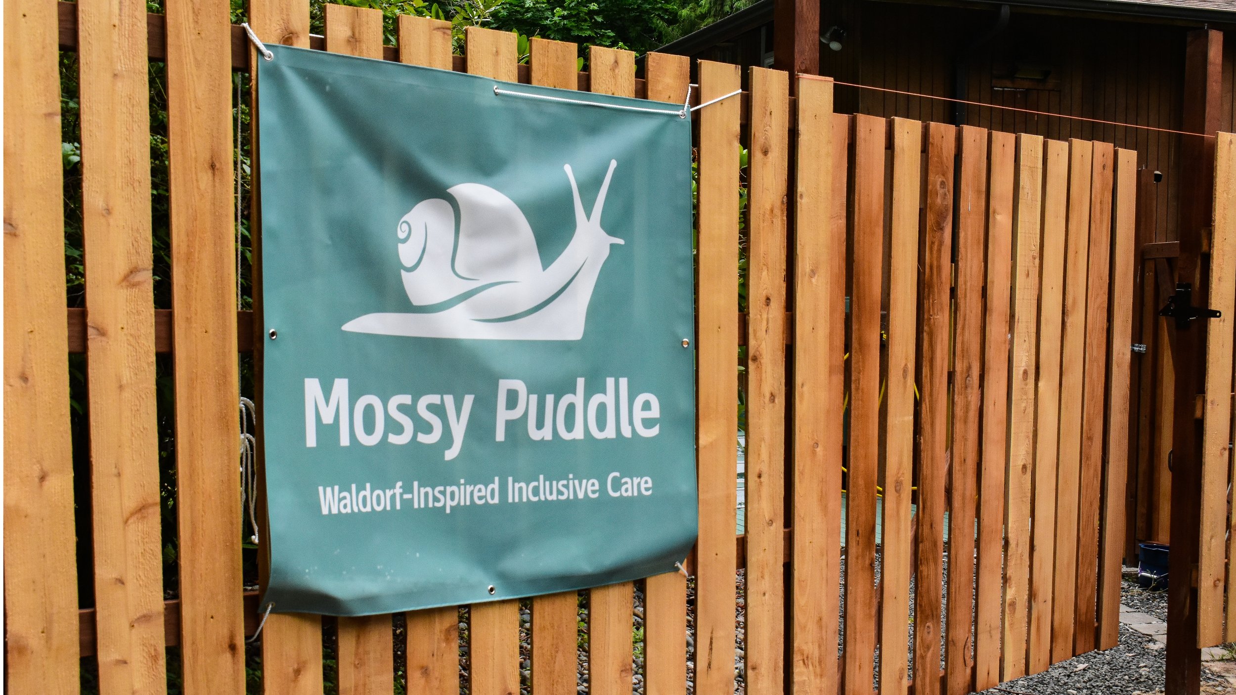 A teal banner attached to a wooden fence, featuring a white graphic of a snail and the text 'Mossy Puddle' and 'Waldorf-Inspired Inclusive Care'.