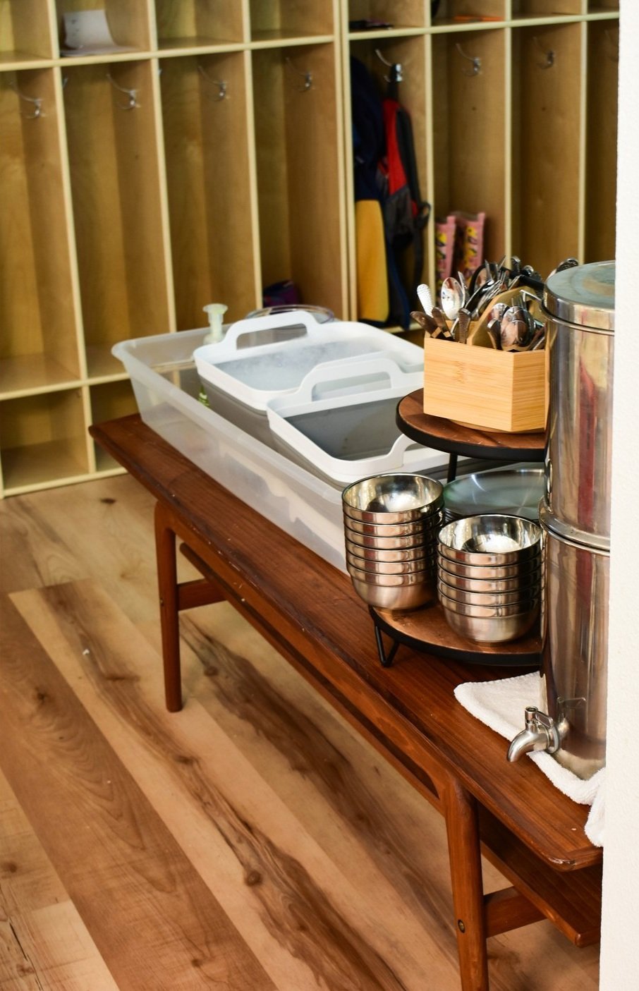 A wooden table holding a clear plastic container with white plastic lids, a small pump bottle, a metal water dispenser with stacked metal bowls on a wooden rack, and a wooden box filled with cutlery in a room with open cubby storage in the background