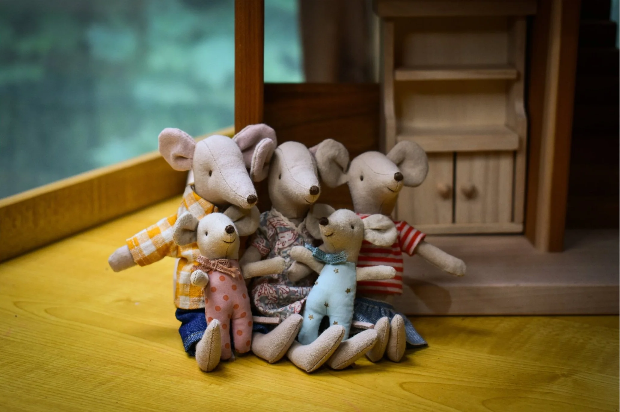 Group of five handmade fabric mice dolls sitting on a wooden surface near a window with a blurry outdoor view, with a wooden shelf in the background.
