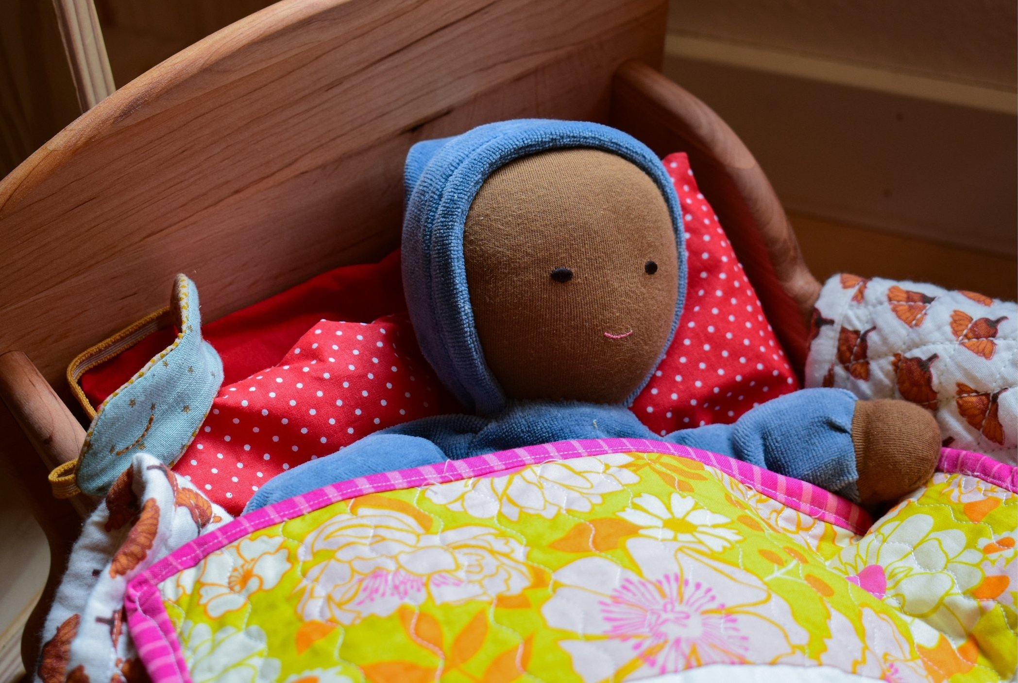 A stuffed doll with a brown face, blue hood, and dark blue outfit sleeping in a bed with a wooden headboard, red polka dot blanket, and a colorful floral quilt.
