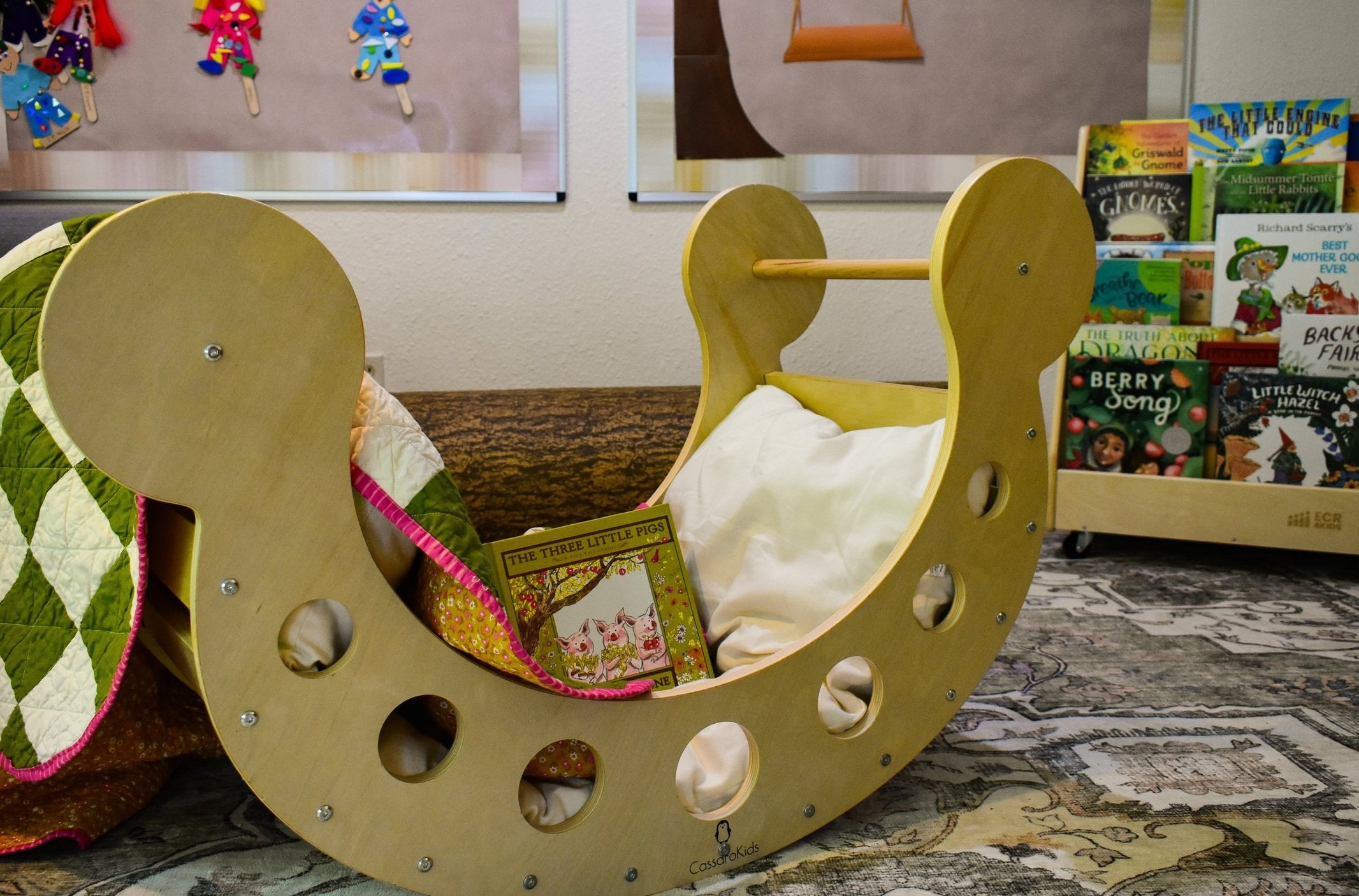 A wooden rocking cradle in a toy store, with a quilted blanket and a children's book inside. Books are displayed on shelves in the background.