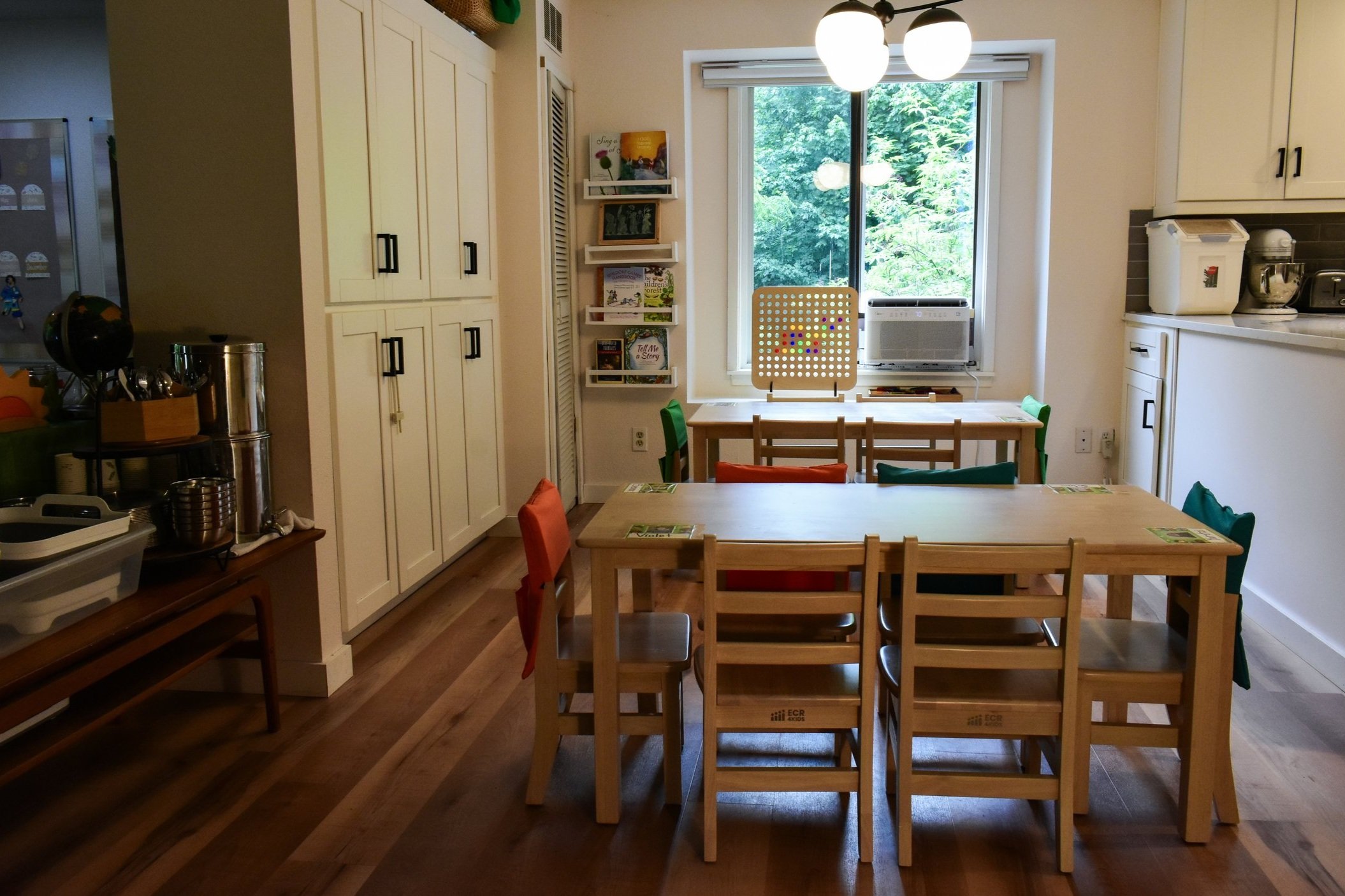A cozy kitchen with wooden tables and colorful chairs near a large window. The room has white cabinets, a bookshelf with books, and various kitchen appliances. There is a small table with utensils and containers on the left side.