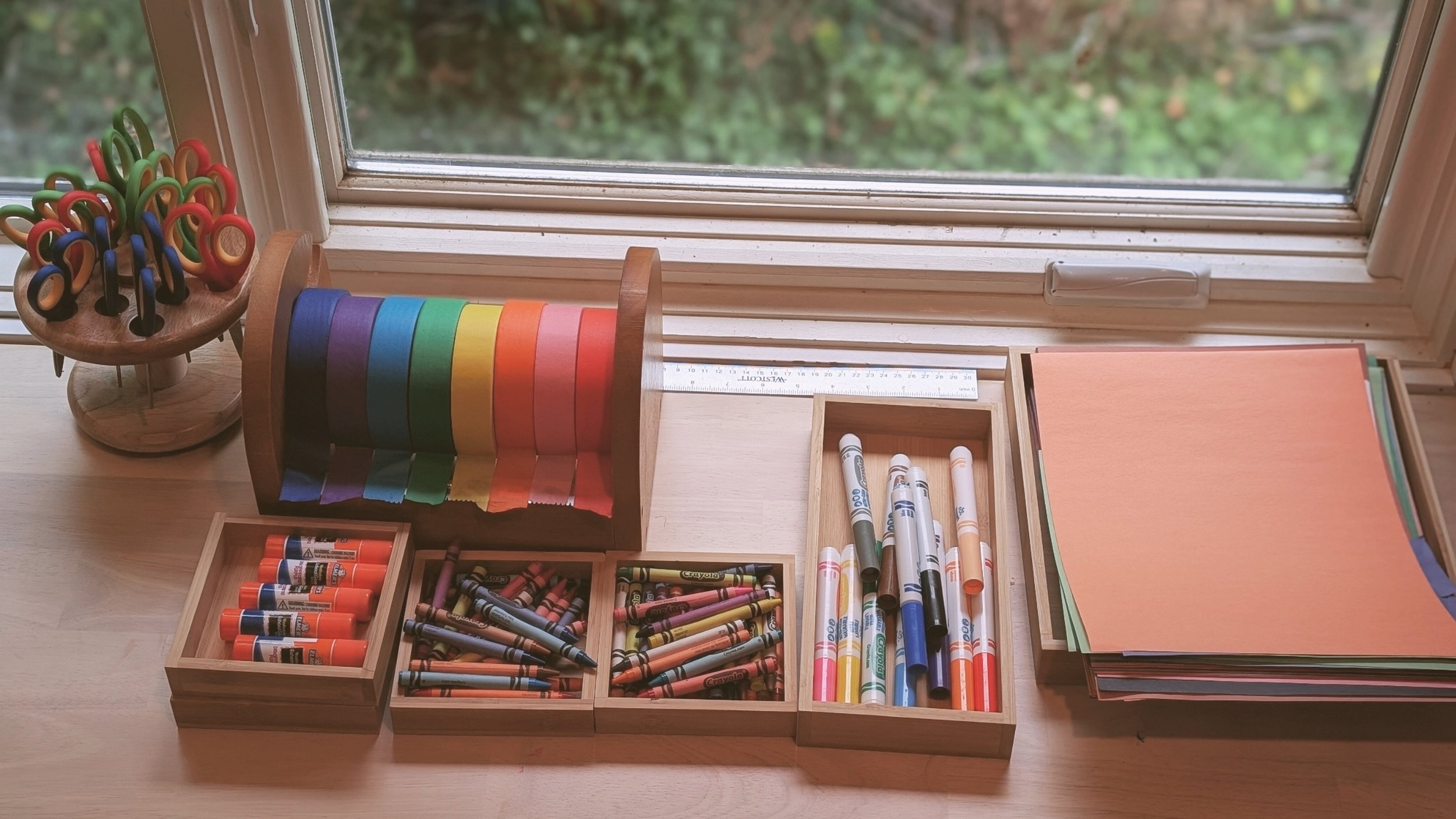Various colored craft supplies arranged on a wooden table near a window, including crayon boxes, glue sticks, markers, colored paper, and a ring of multicolored tape.