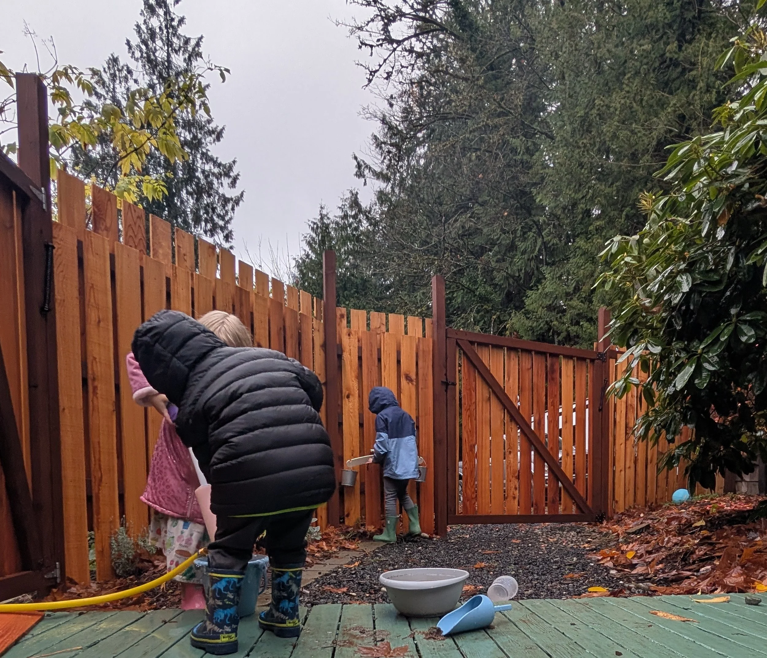 Children playing outdoors during rainy weather, washing toys and playing near a wooden fence, with trees and cloudy sky in the background.