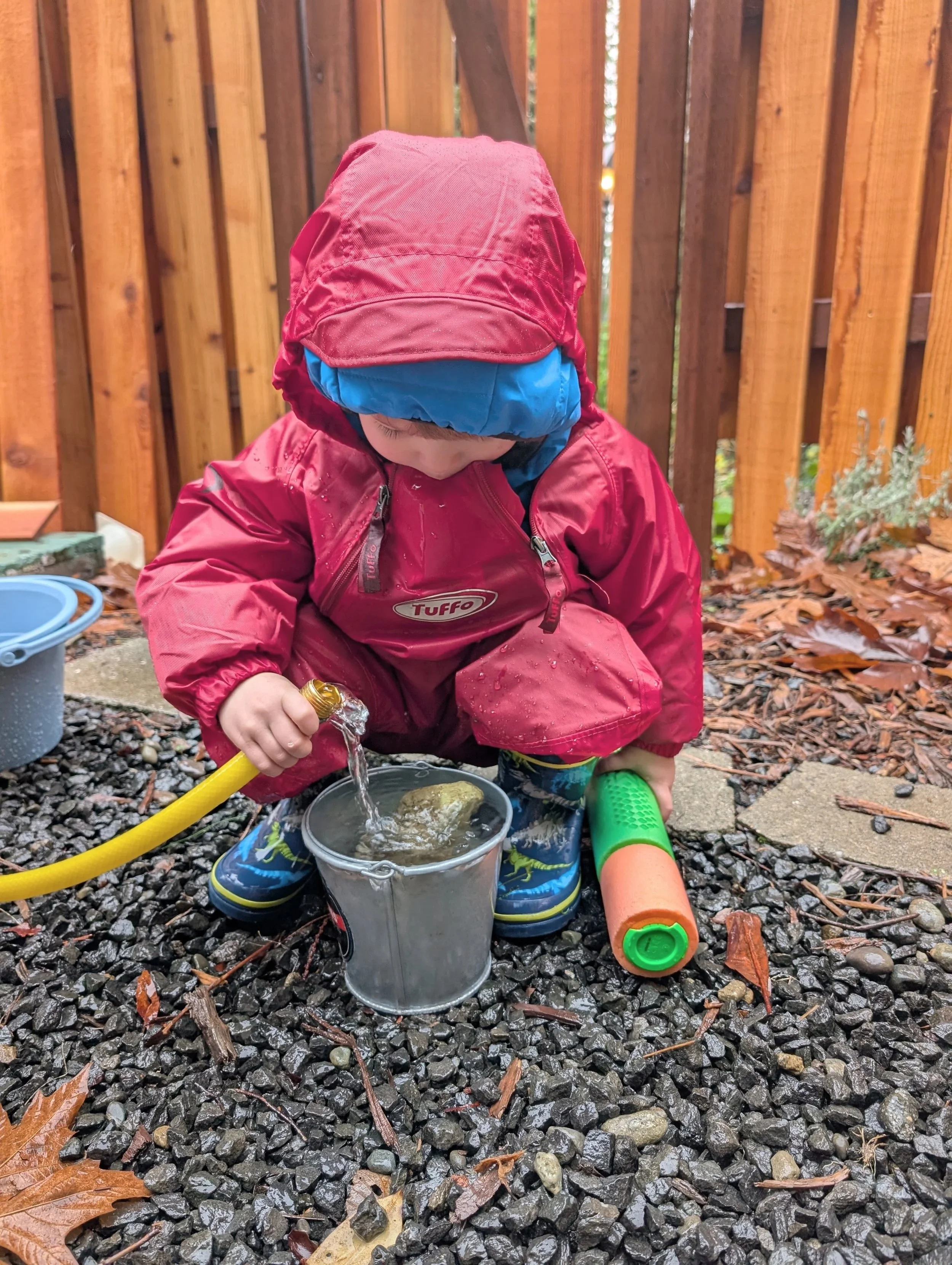 A young child dressed in a red waterproof jacket and blue boots playing outside on gravel, filling a small metal bucket with water from a yellow hose, with a plastic toy around them and a wooden fence in the background.