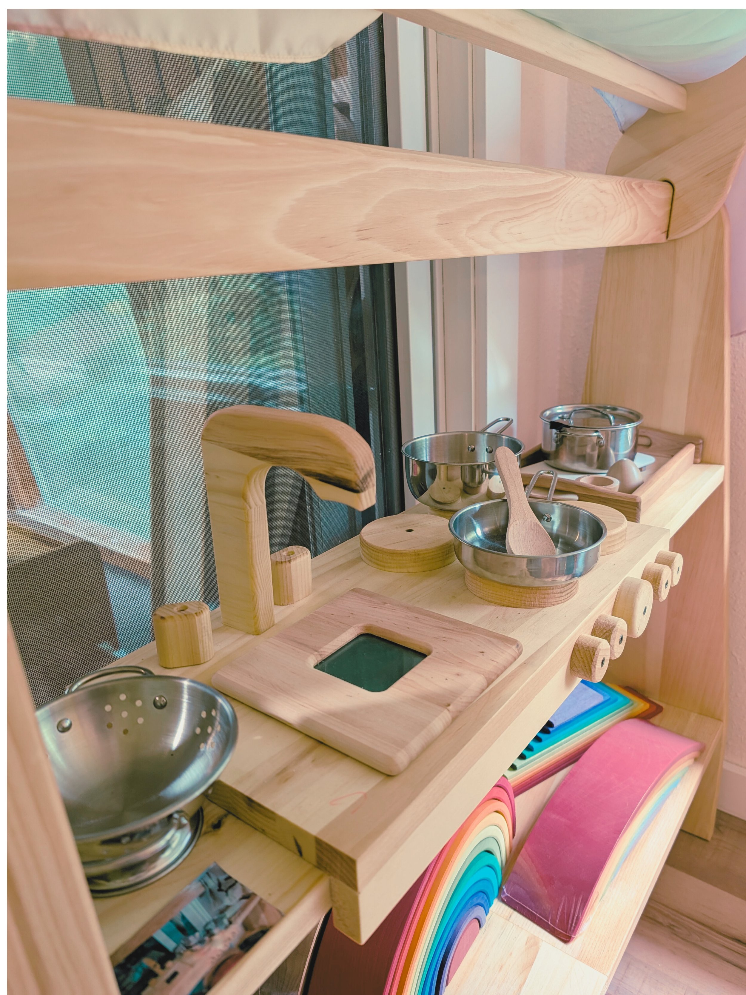 Wooden play kitchen set with metal pots, pans, and utensils, colorful rainbow-themed decor, and a chalkboard surface, positioned near a window.