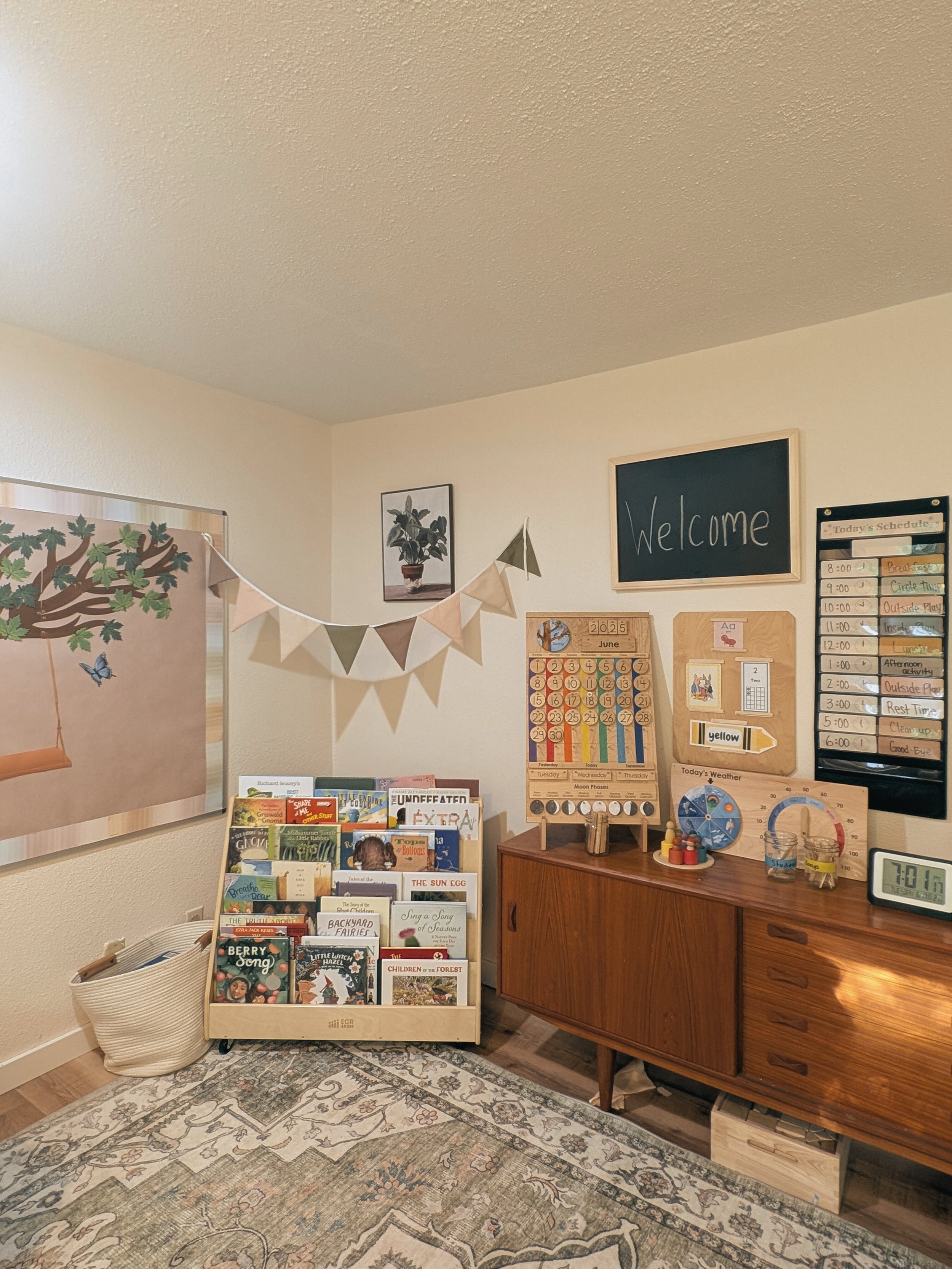 A children's classroom with a wooden bookshelf filled with children's books, a chalkboard with a welcome message, a weekly schedule chart, weather and moon phase charts, and decorative bunting flags on the wall.