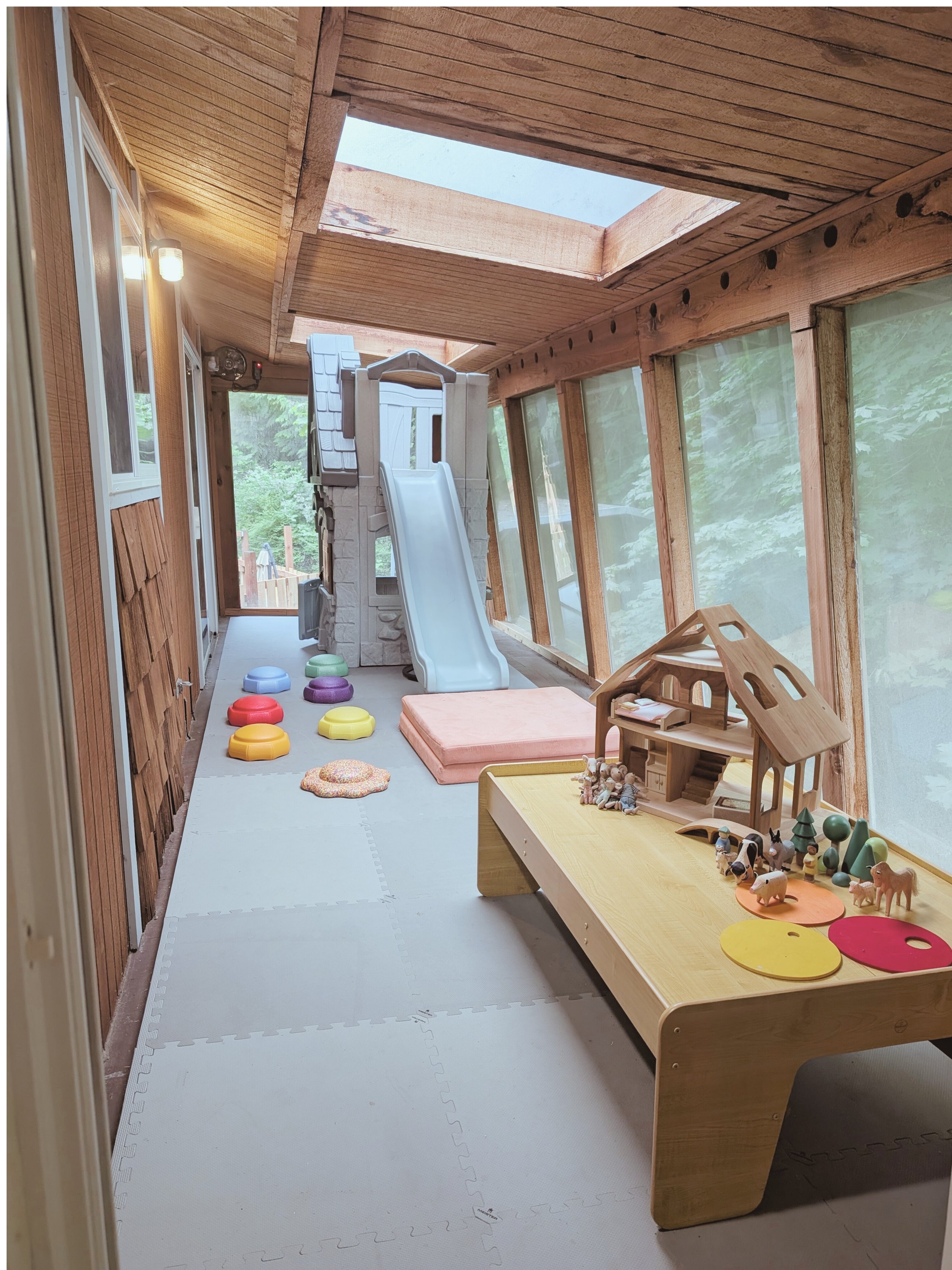 Enclosed porch with wooden walls and ceiling, large windows, a play slide, colorful stepping stones, a dollhouse, and plush mats.