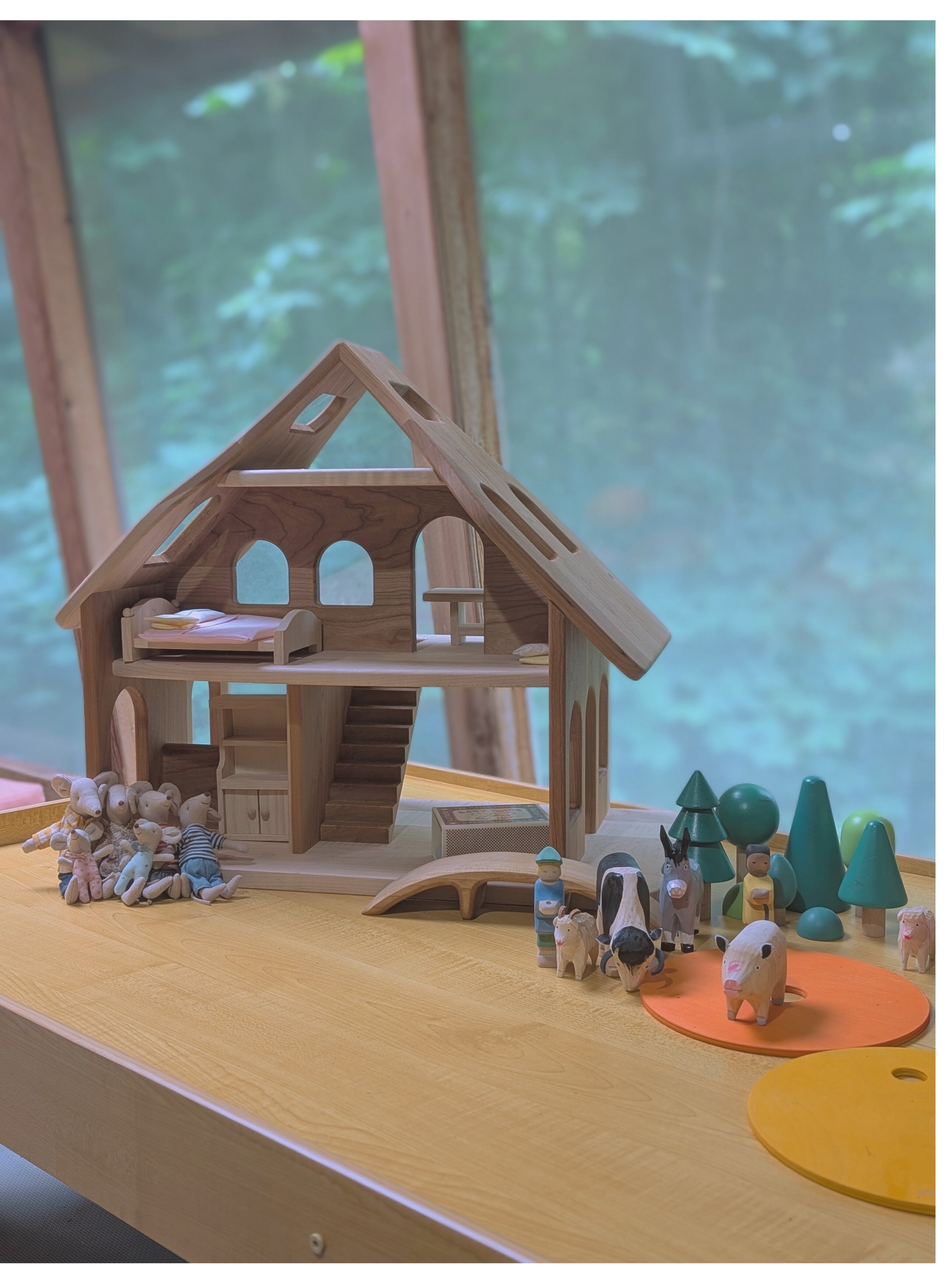 Wooden dollhouse on a table with toy animals, trees, and figures surrounding it, with a light blue background.