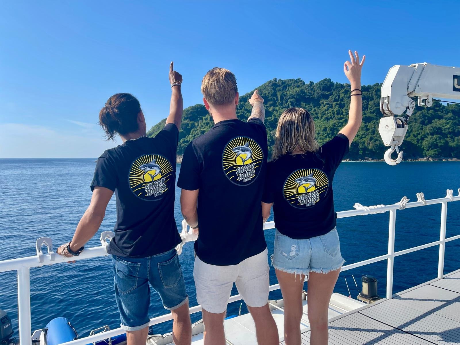 Tre giovani indossano magliette con il logo 'Shark House' su una nave, guardando un paesaggio marino con una collina verde e un cielo sereno.