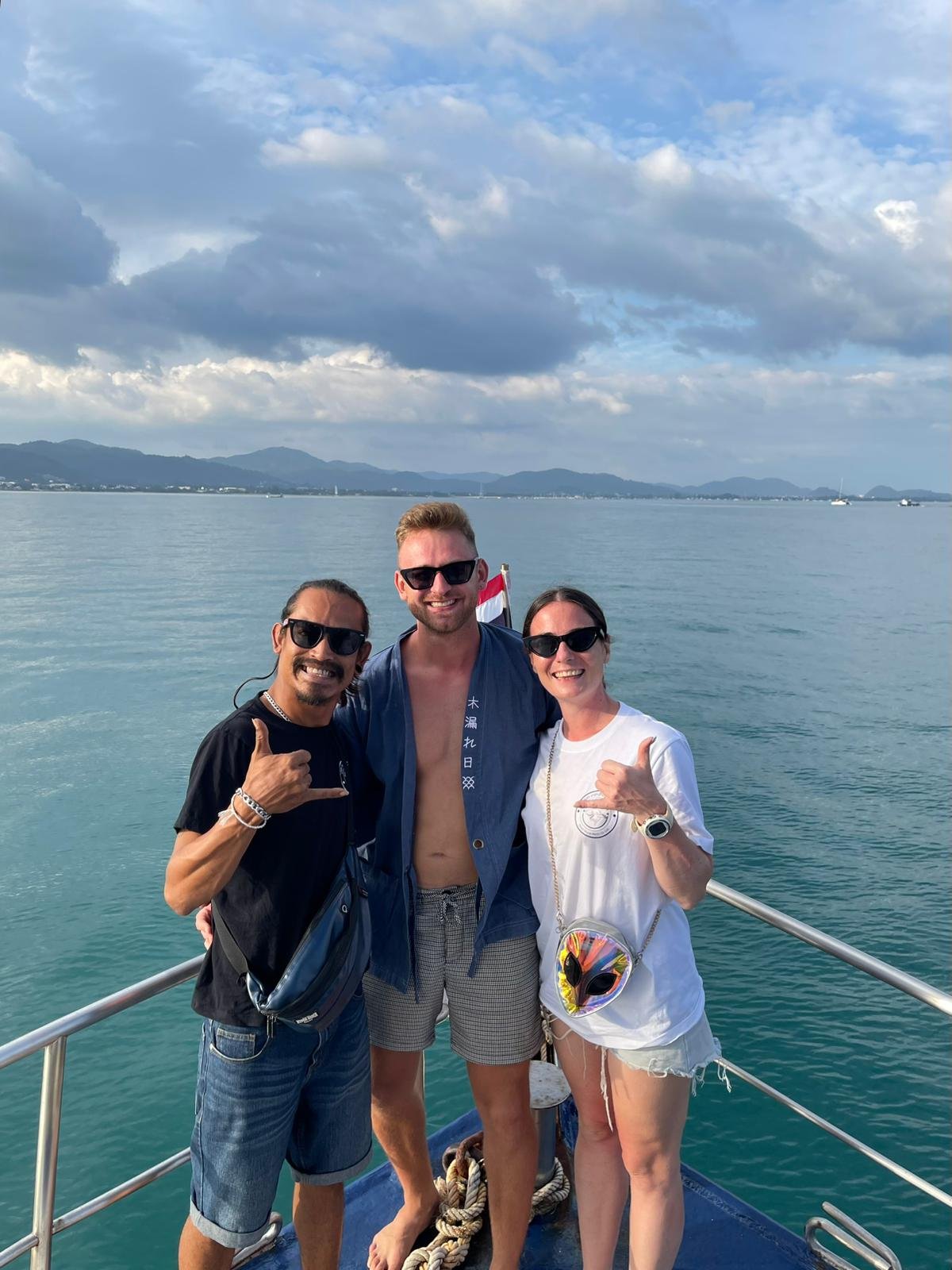 Tre persone sorridenti su una barca sul mare, due uomini e una donna, tutti con occhiali da sole, con sfondo di cielo e montagne lontane.