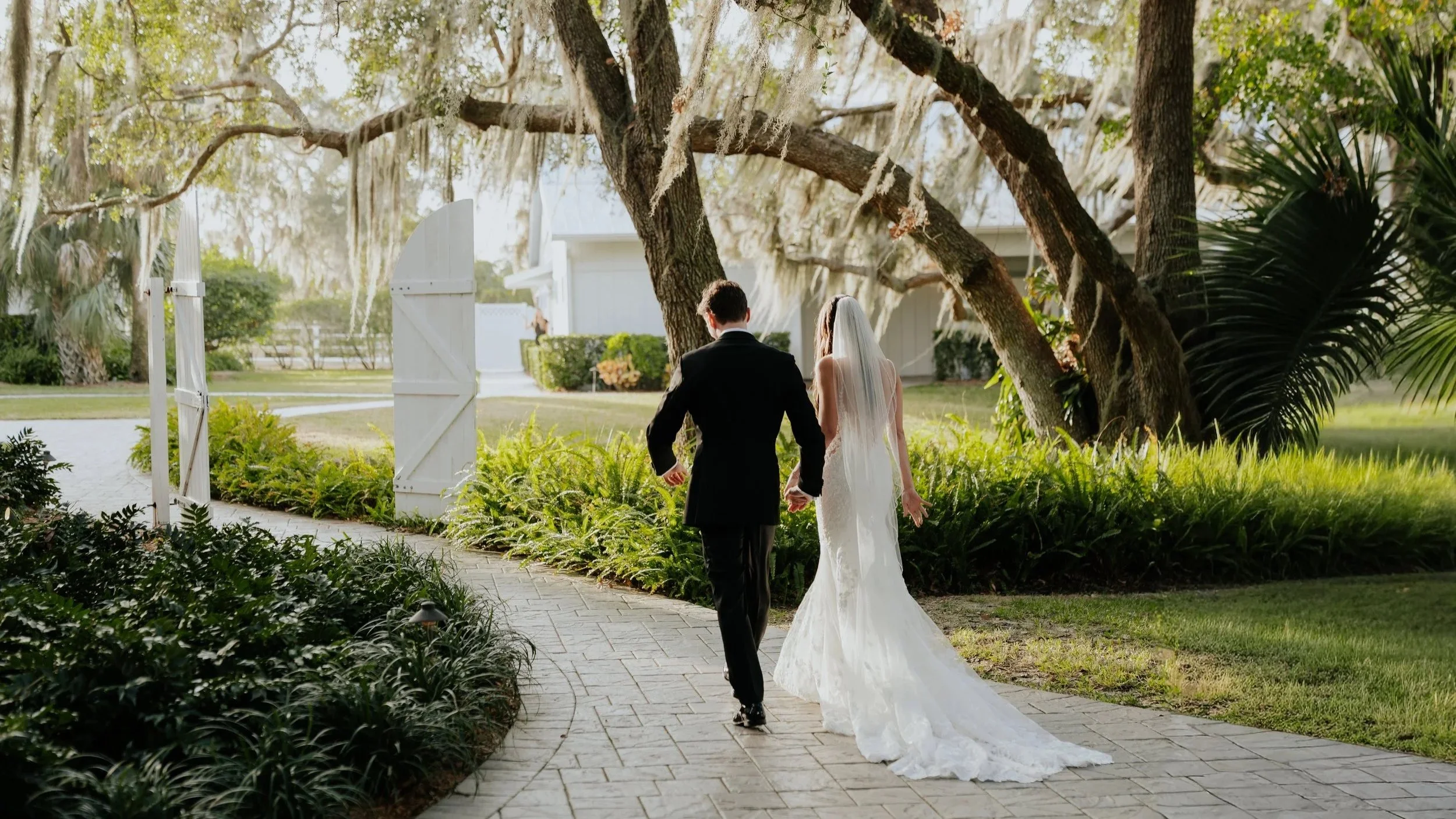 A bride and groom walking hand in hand on a stone pathway in a garden, with trees and greenery in the background, during daytime.