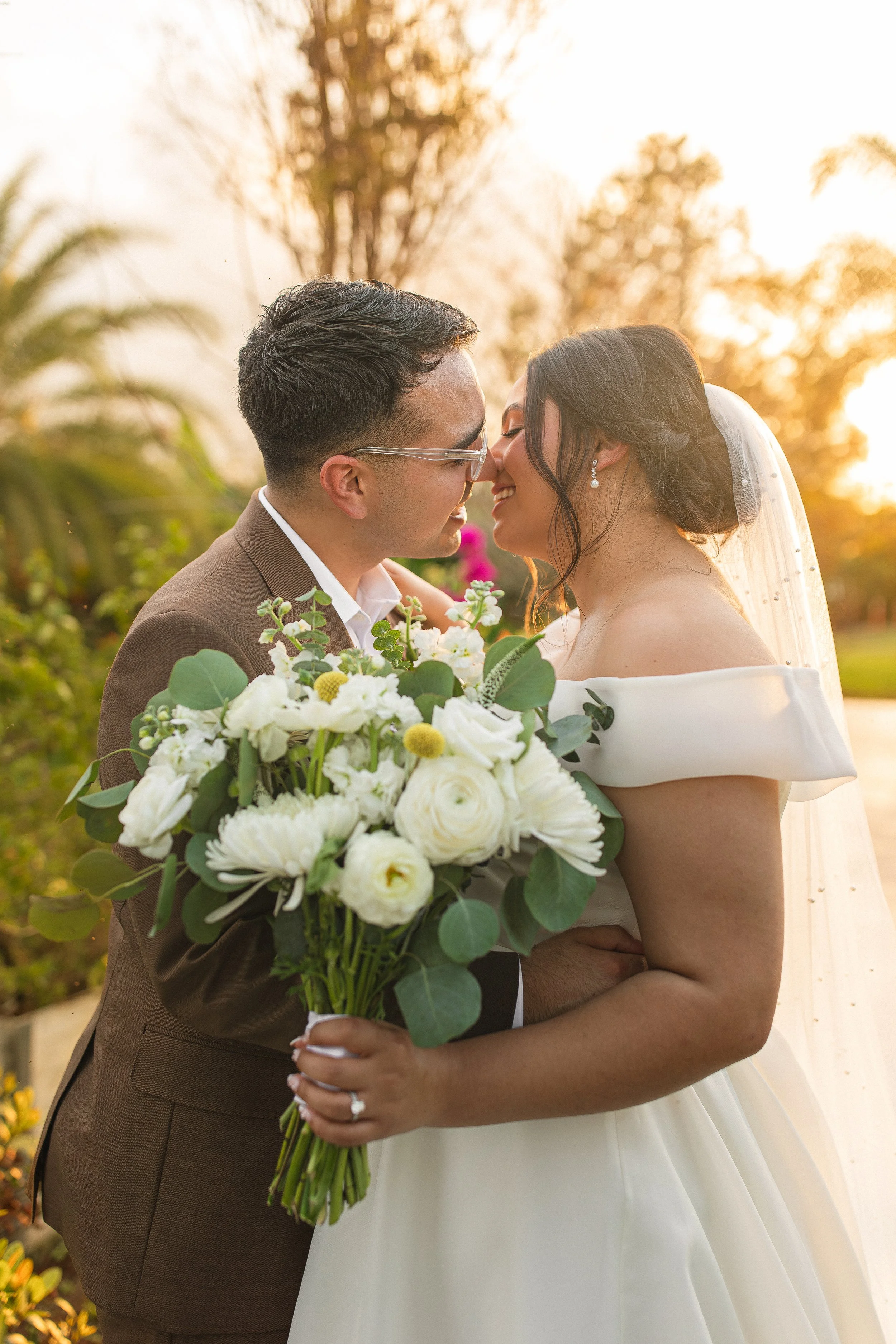 Bride and groom smiling and holding a bouquet, about to kiss, in a garden setting.