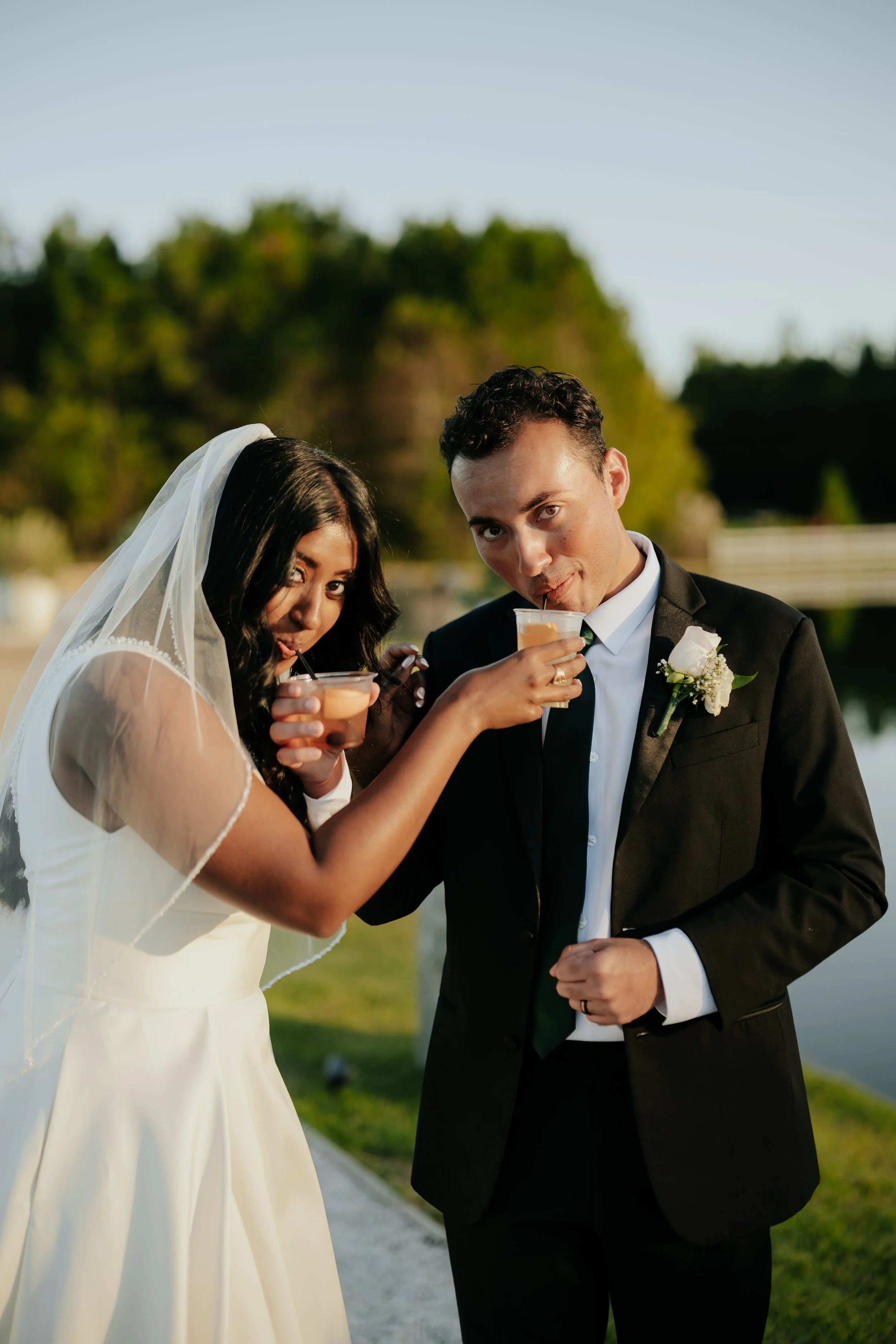 A bride and groom in wedding attire sharing drinks outdoors near a body of water during sunset.