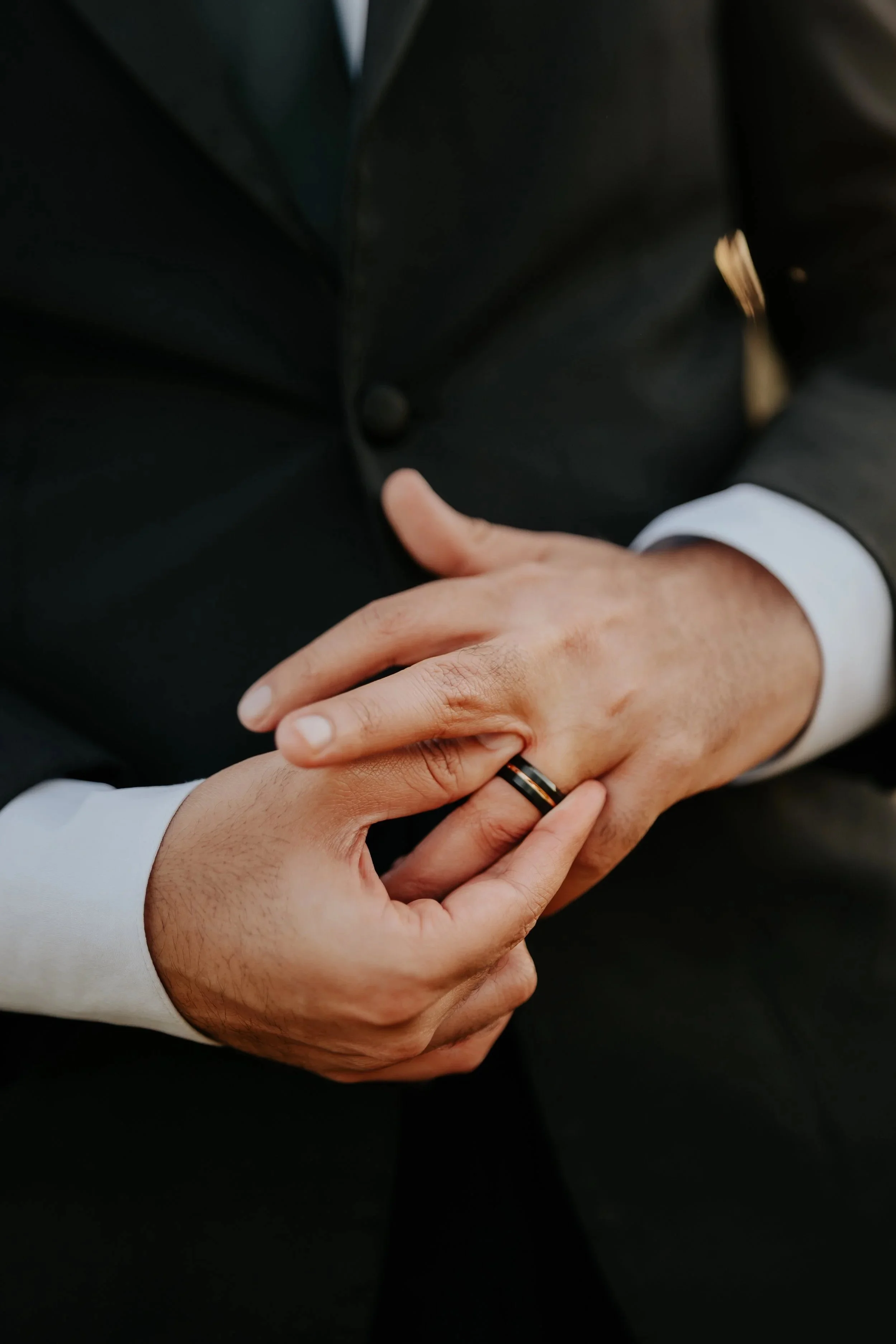 Close-up of wedding rings being held by two people, one wearing a black suit and the other a white shirt, symbolizing a wedding or commitment.