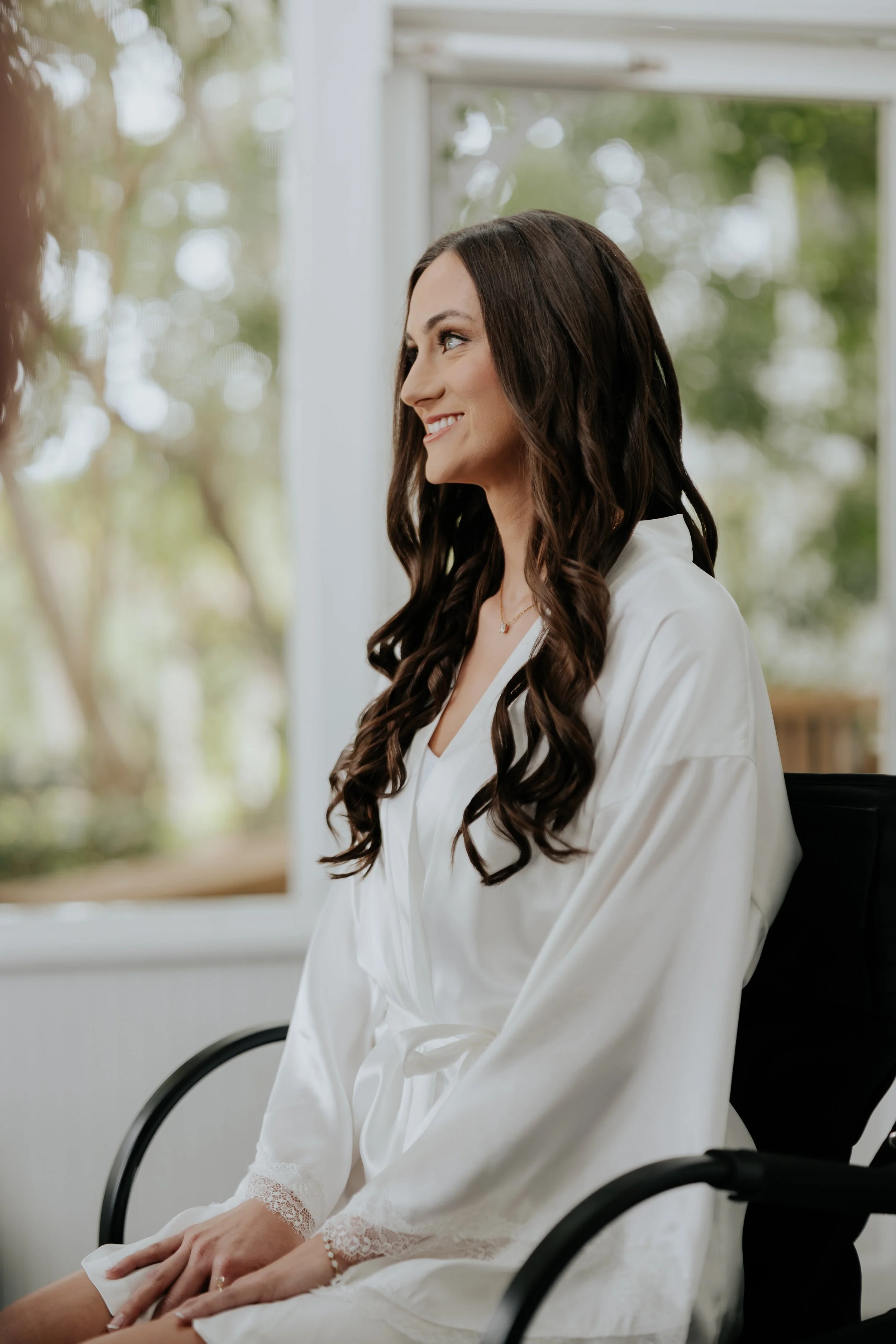 A woman with long, wavy dark hair, smiling softly, wearing a white satin robe with lace trim, sitting by a window with greenery outside.