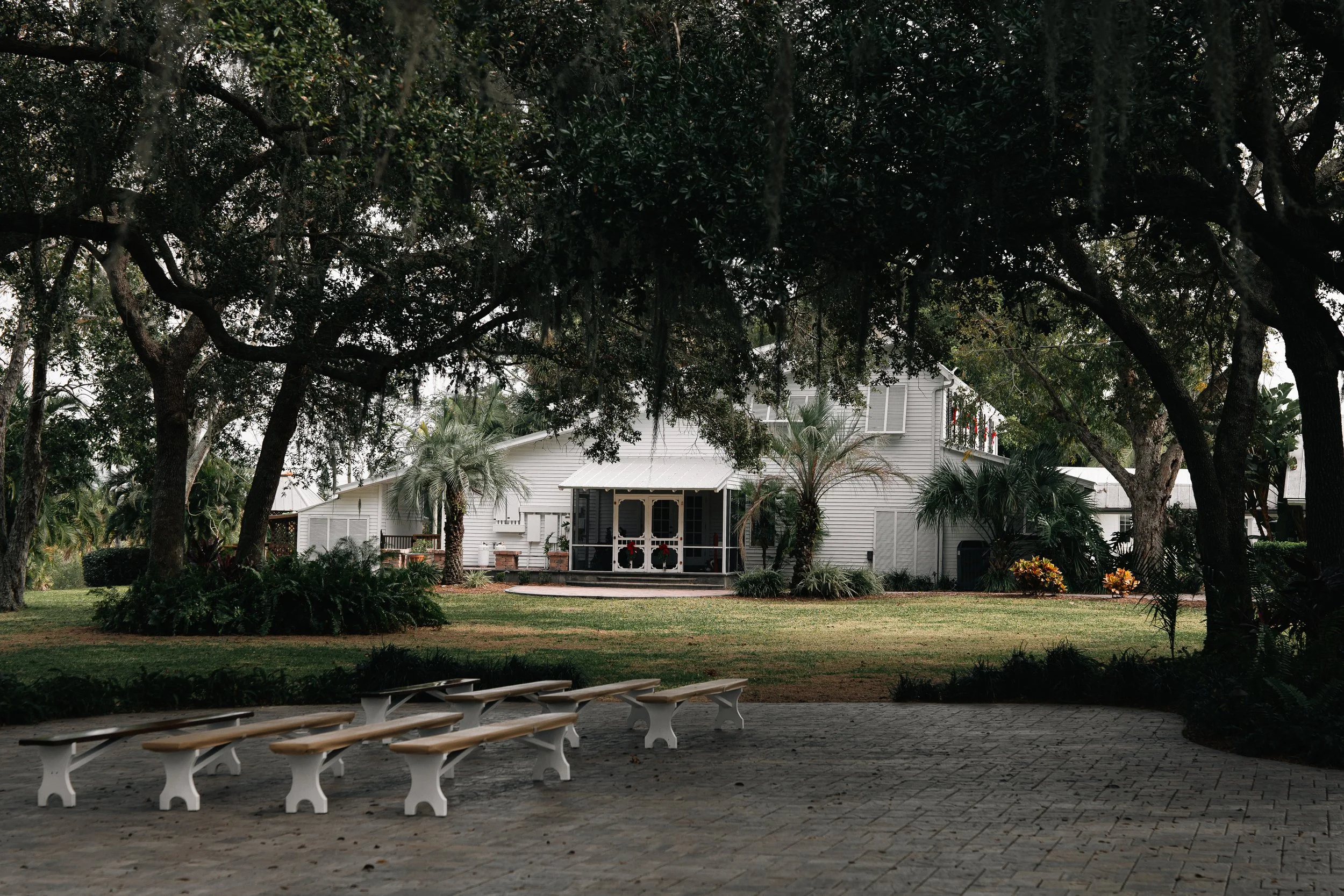 White barn-style building surrounded by trees and palm plants, with rows of white benches on a paved area in the foreground.