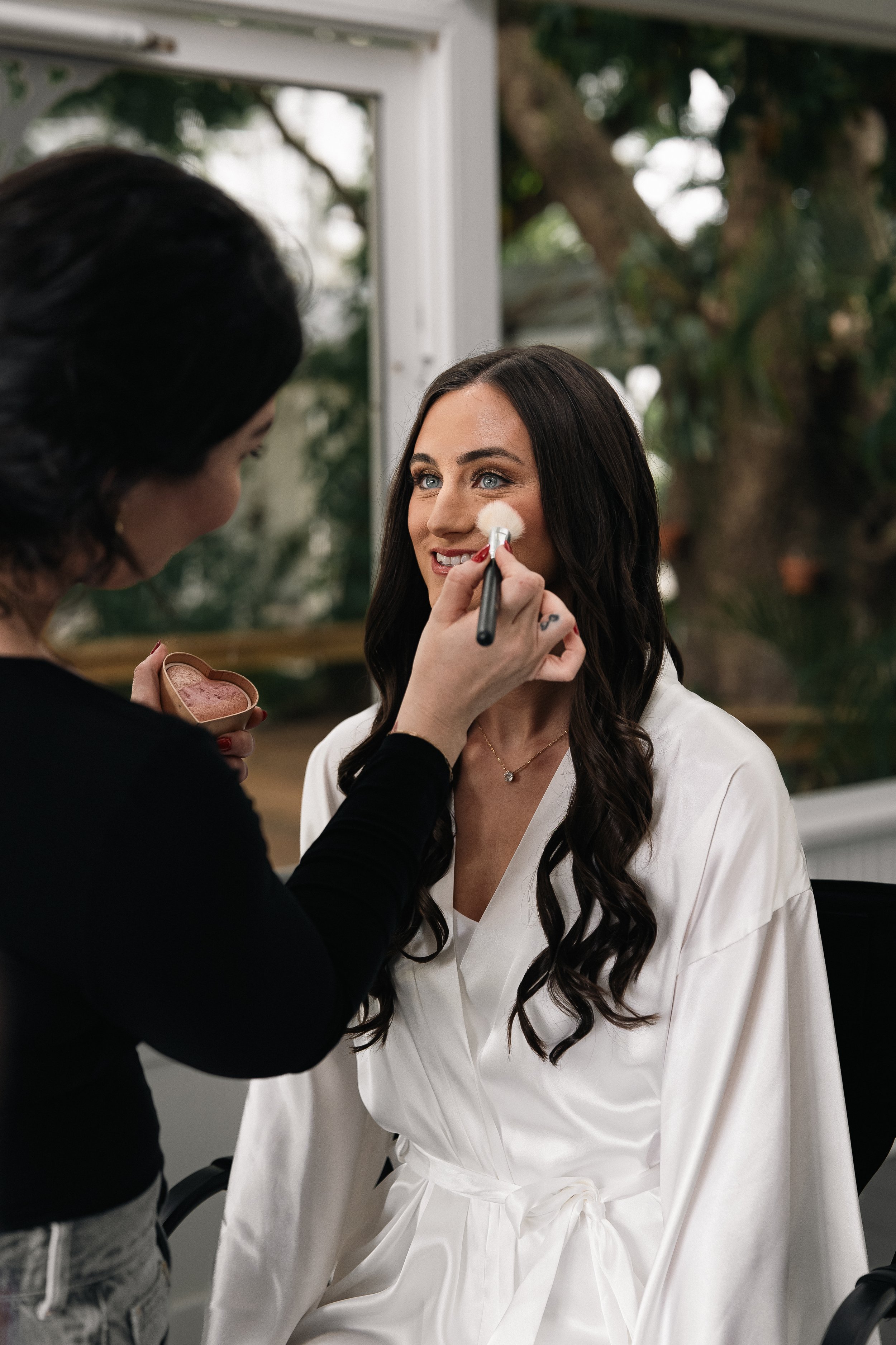 A woman applying makeup to another woman's face with a brush in a well-lit room, while the seated woman wears a white robe.