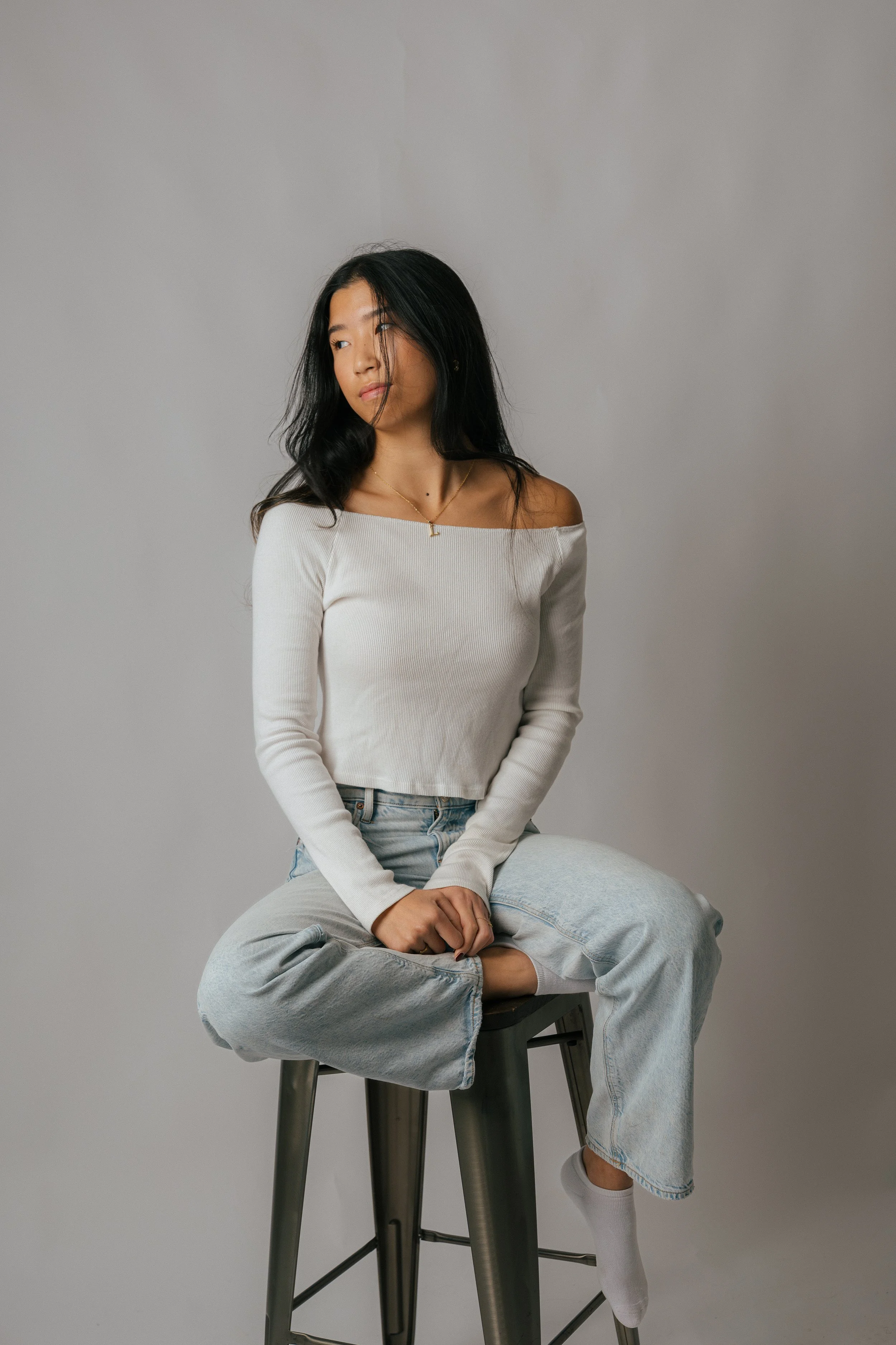 Person in off-shoulder white top and jeans seated on stool