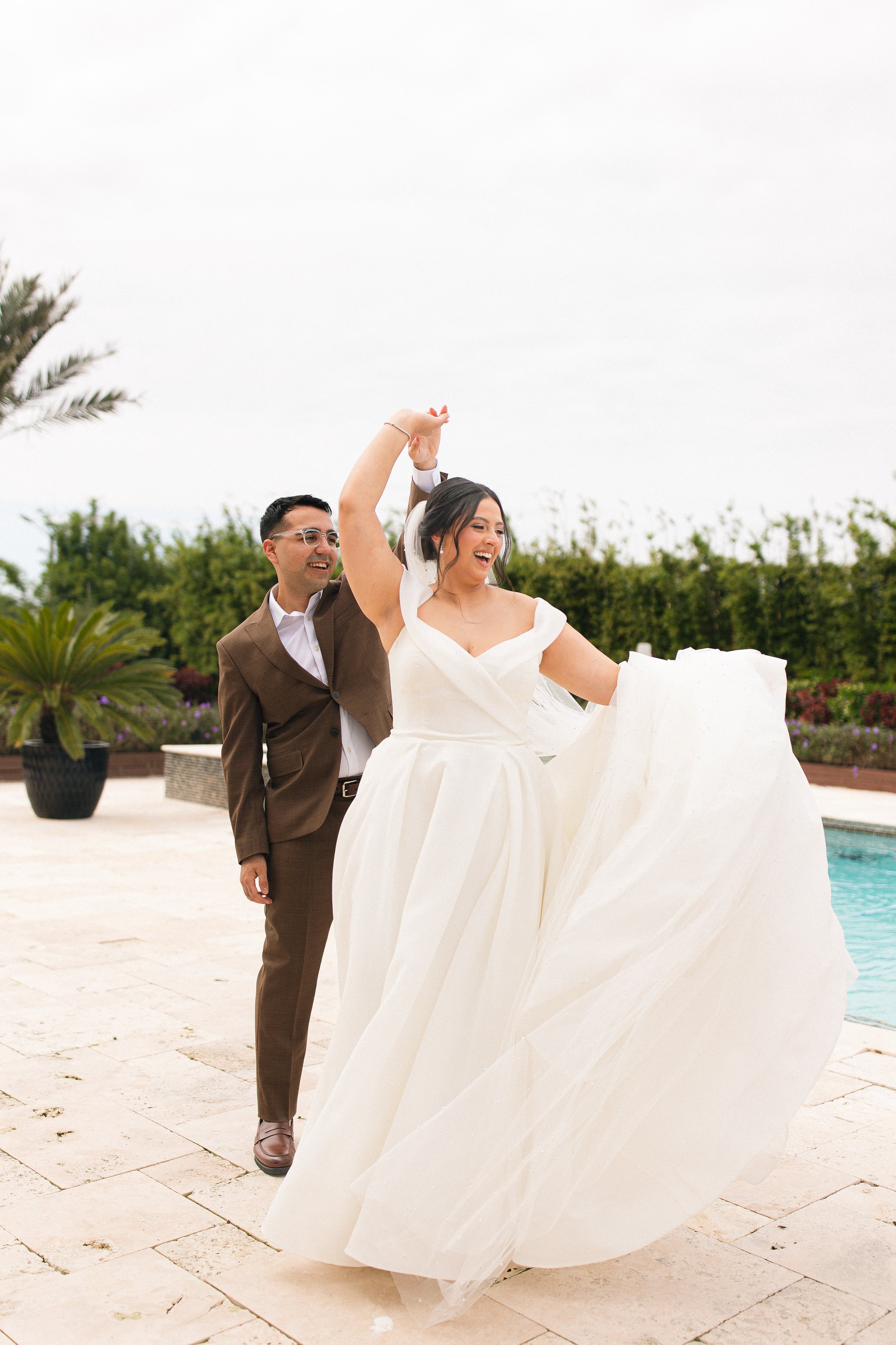 A couple dancing outdoors by a pool, with the woman in a white wedding dress and the man in a brown suit, surrounded by greenery.