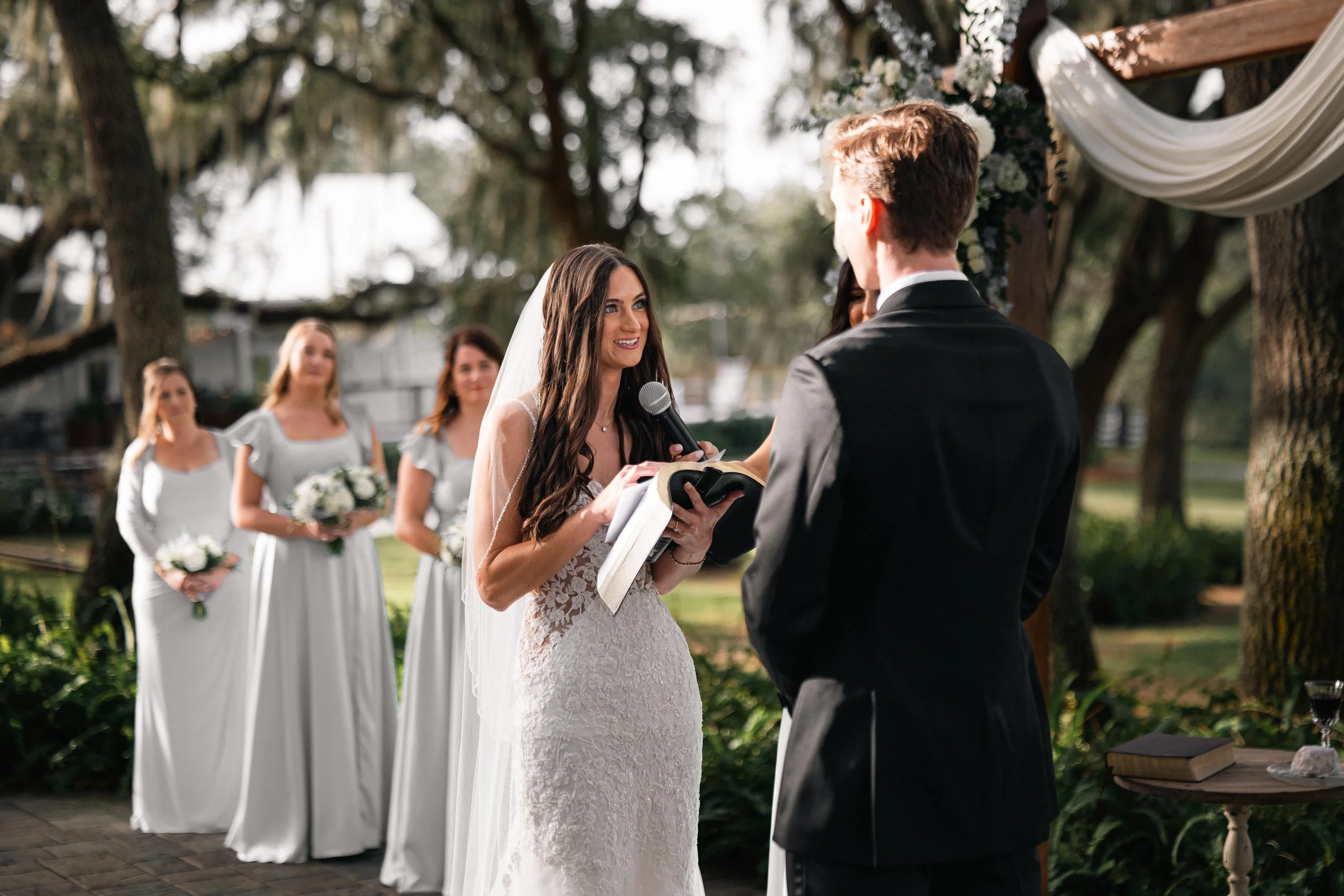 Bride reading vows to groom at outdoor wedding ceremony, with bridesmaids in background holding bouquets.
