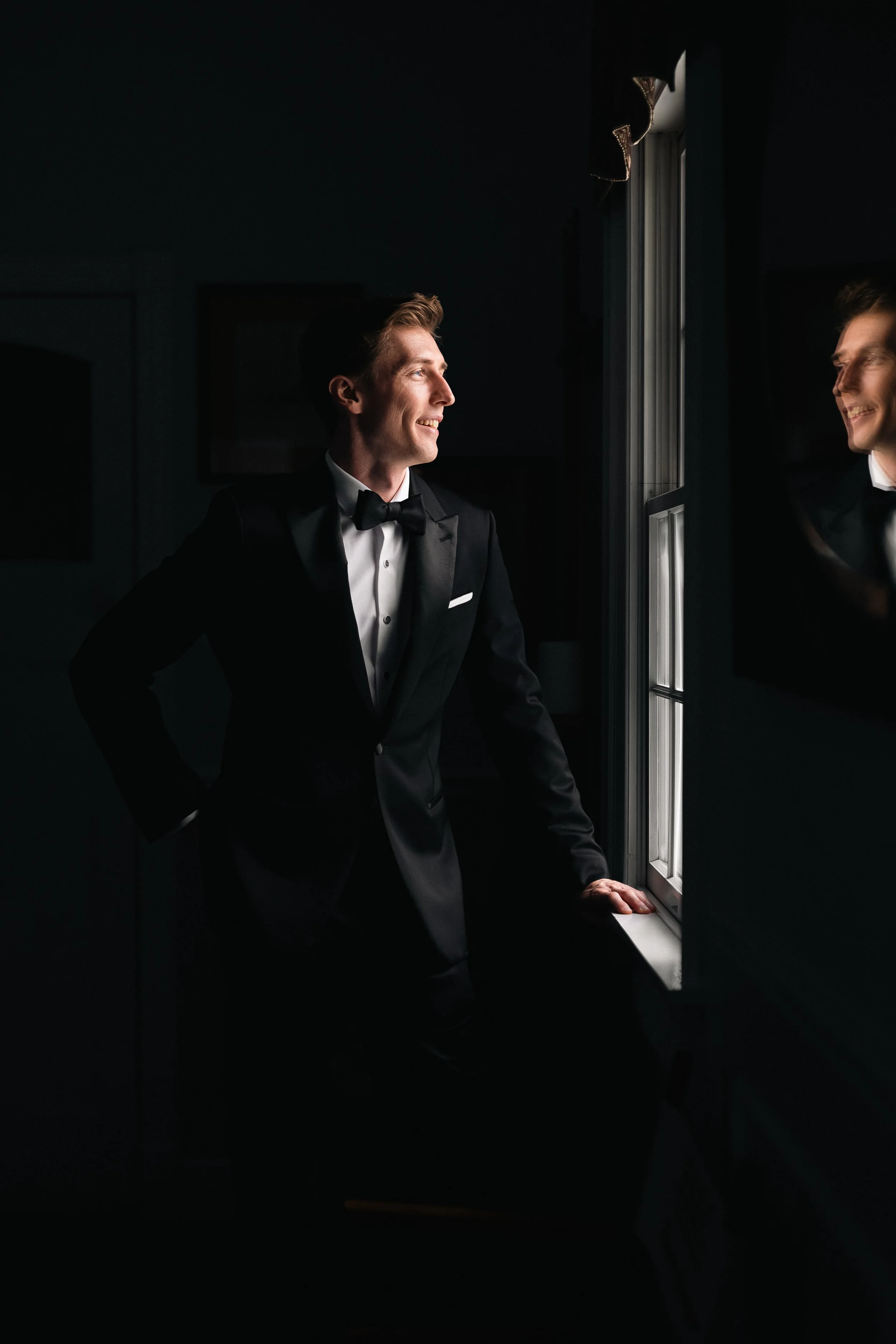Man in a tuxedo standing by a window, smiling at his reflection in the glass.