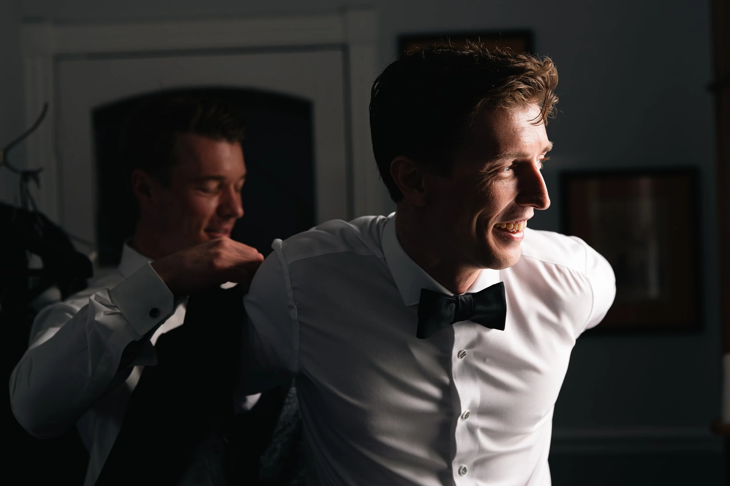 Two men dressing in formal attire, one helping the other with a jacket, both wearing white shirts and black bow ties, in a dimly lit room.