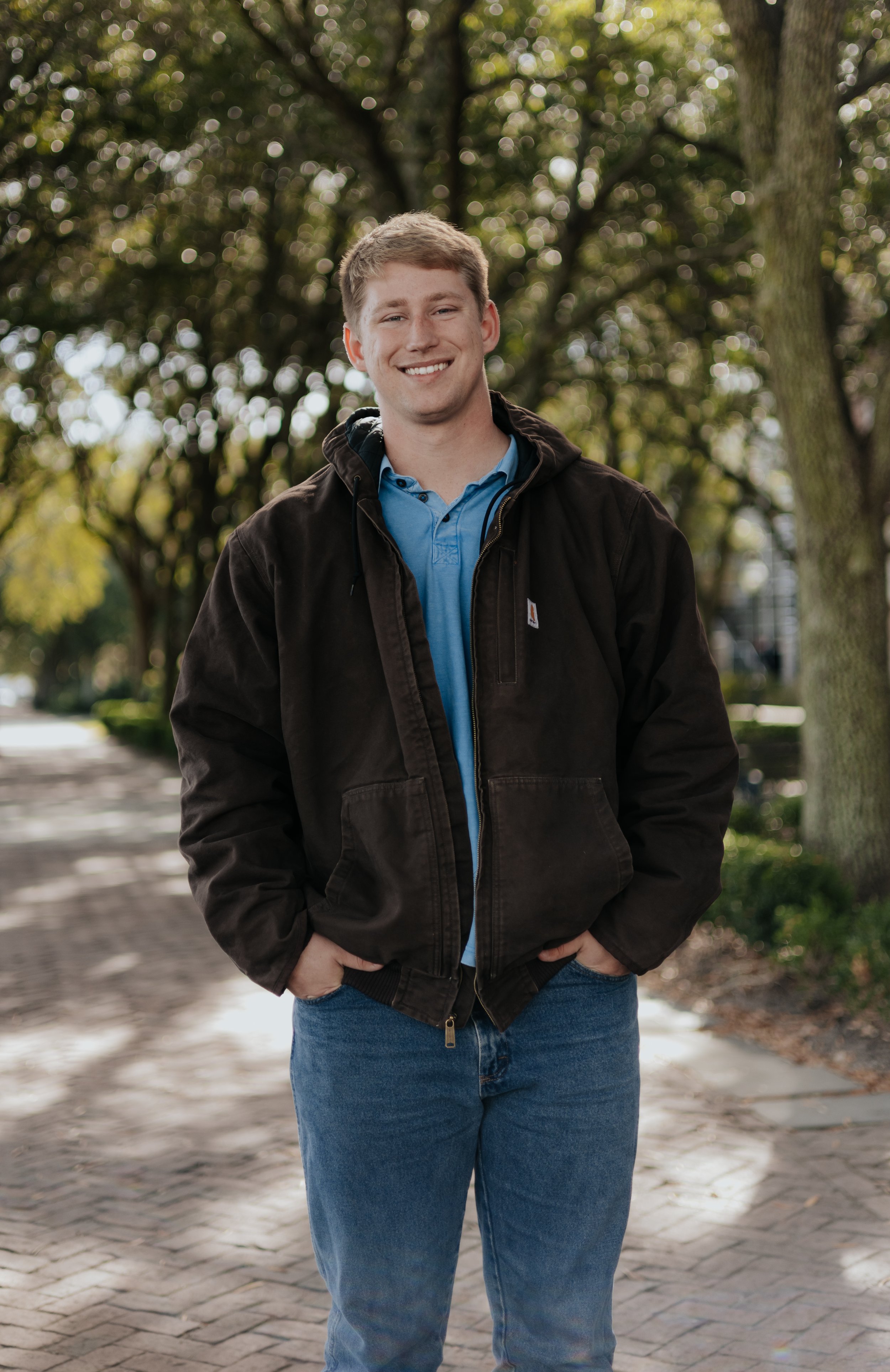 A young man with short blond hair smiling while standing outdoors on a sunny day, wearing a black jacket over a blue polo shirt and blue jeans, with trees and a sidewalk in the background.
