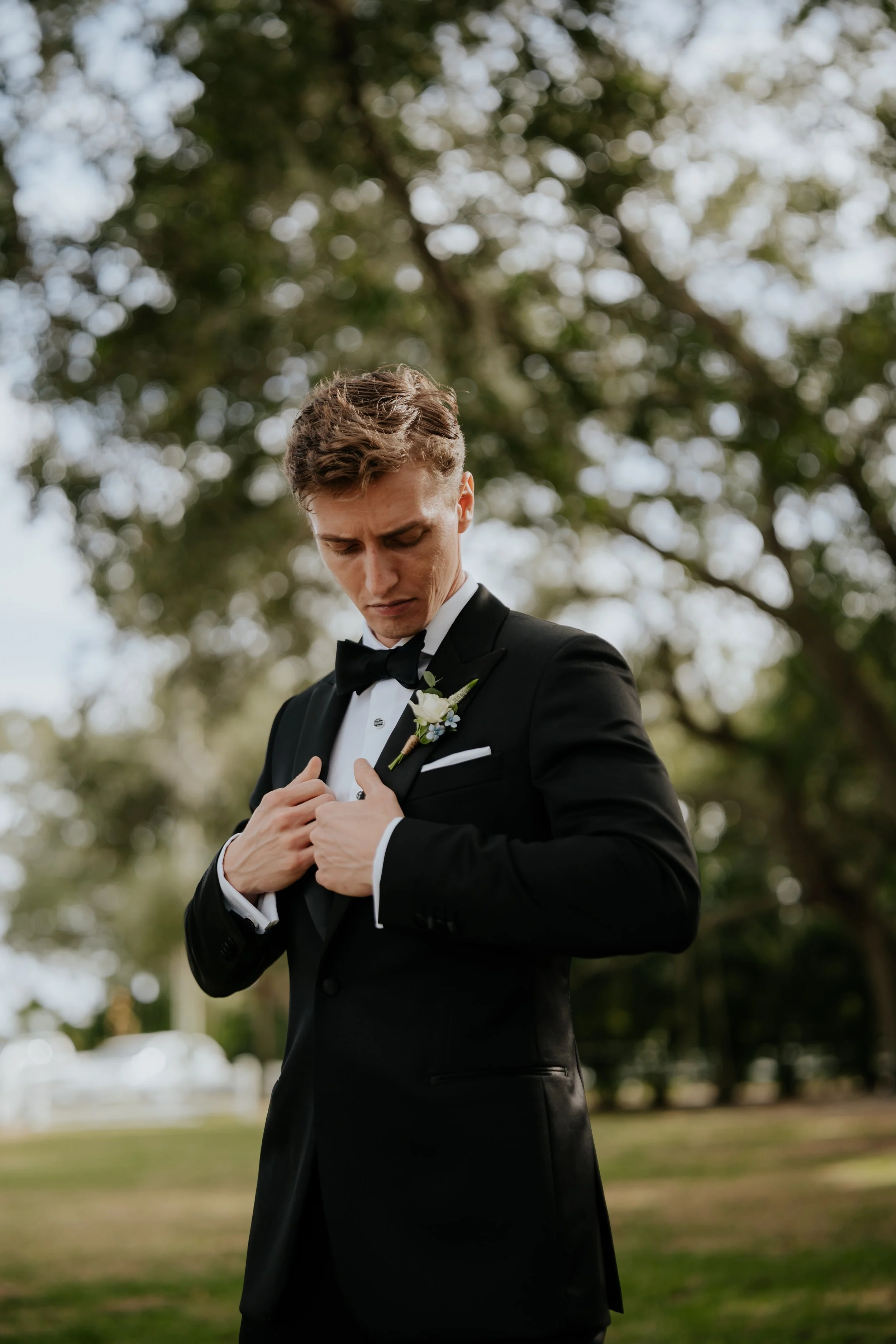 An attractive groom in a black tuxedo with a bow tie and white shirt, adjusting his tuxedo jacket, standing outdoors with trees in the background, on a cloudy day.