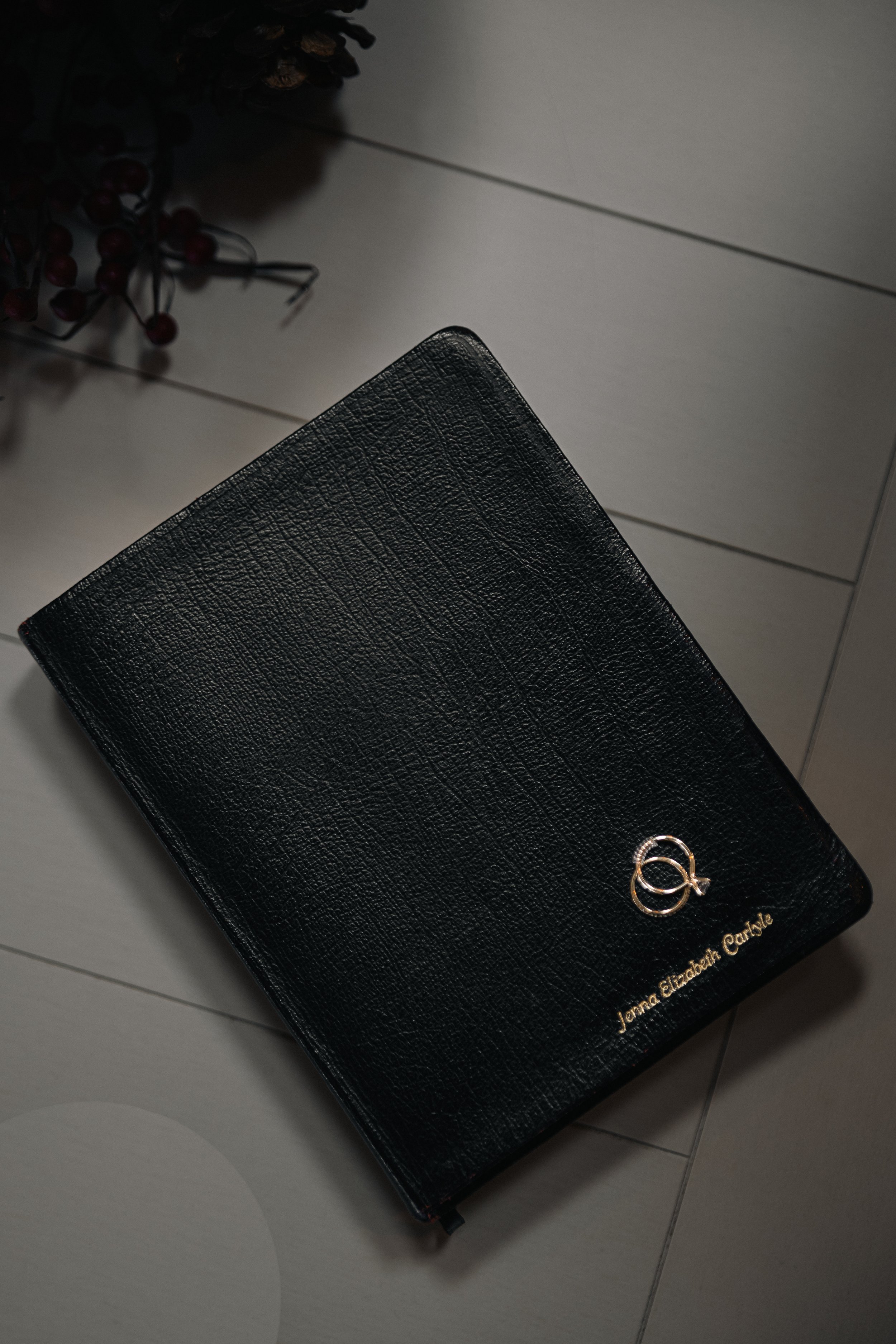 Black leather-bound book with gold initials and name on the cover, placed on a tiled surface with decorative elements nearby.