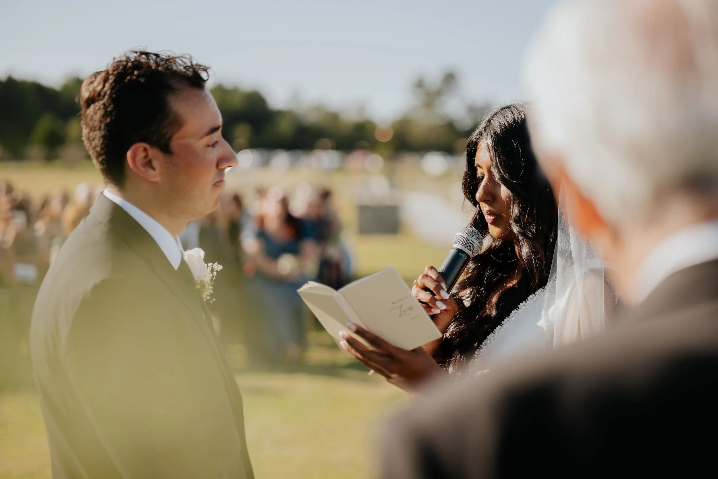 A wedding ceremony outdoors with a bride and groom facing each other, a woman reading vows into a microphone, and guests in the background.