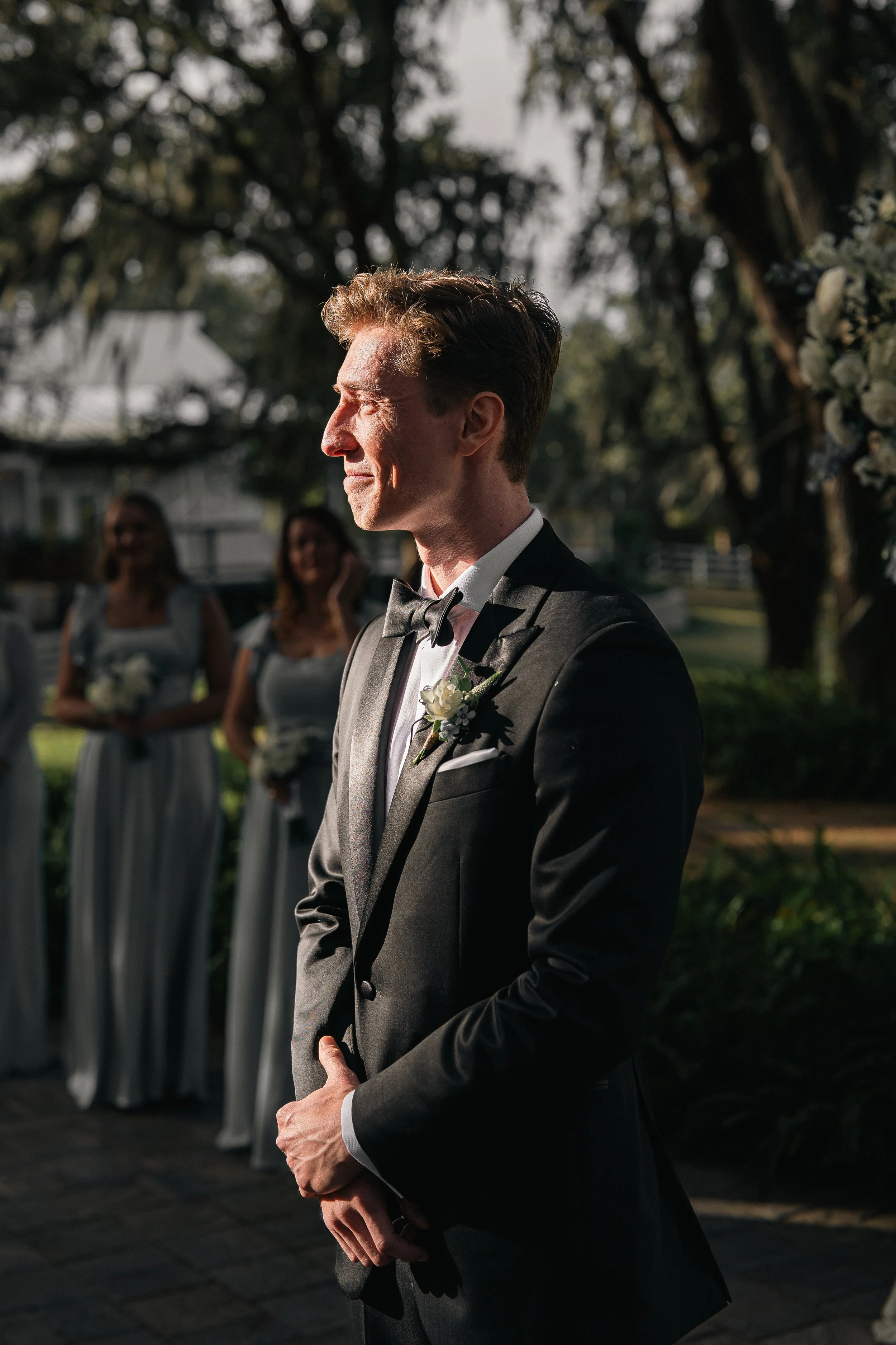 A groom in a black suit and bow tie stands outdoors during a wedding ceremony, holding his hands together, with sunlight on his face and blurred bridesmaids in the background.
