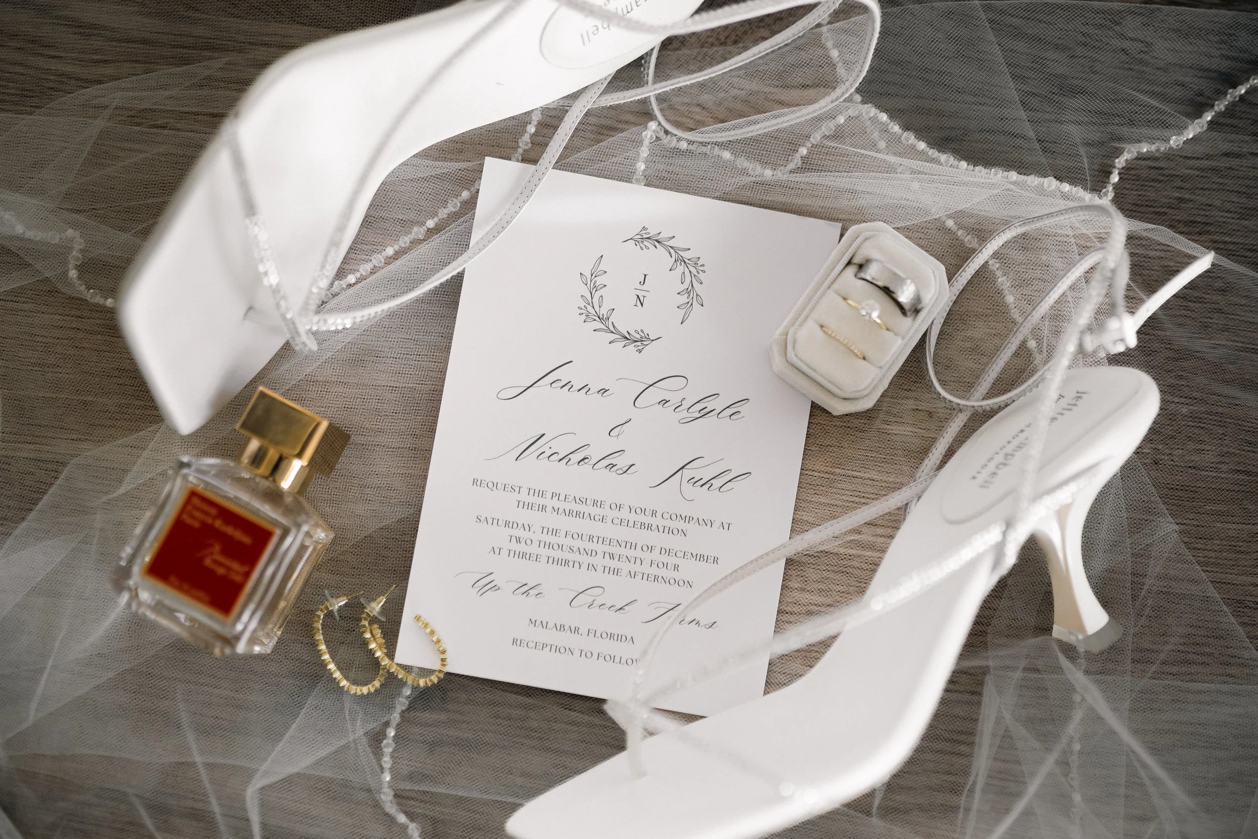 Wedding invitation layout with white high heels, perfume bottle, jewelry, and wedding rings on a veil.