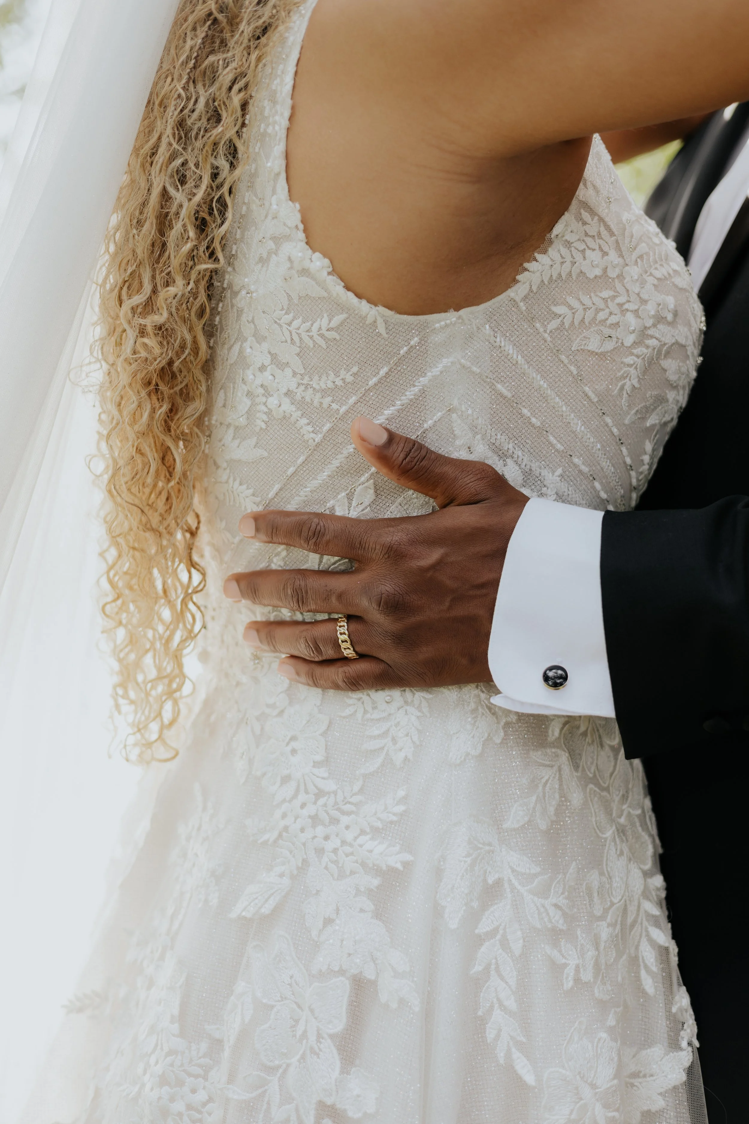 Close-up of a bride wearing an embroidered lace wedding dress and a groom in a tuxedo with a white shirt, with the groom's hand on the bride's chest.