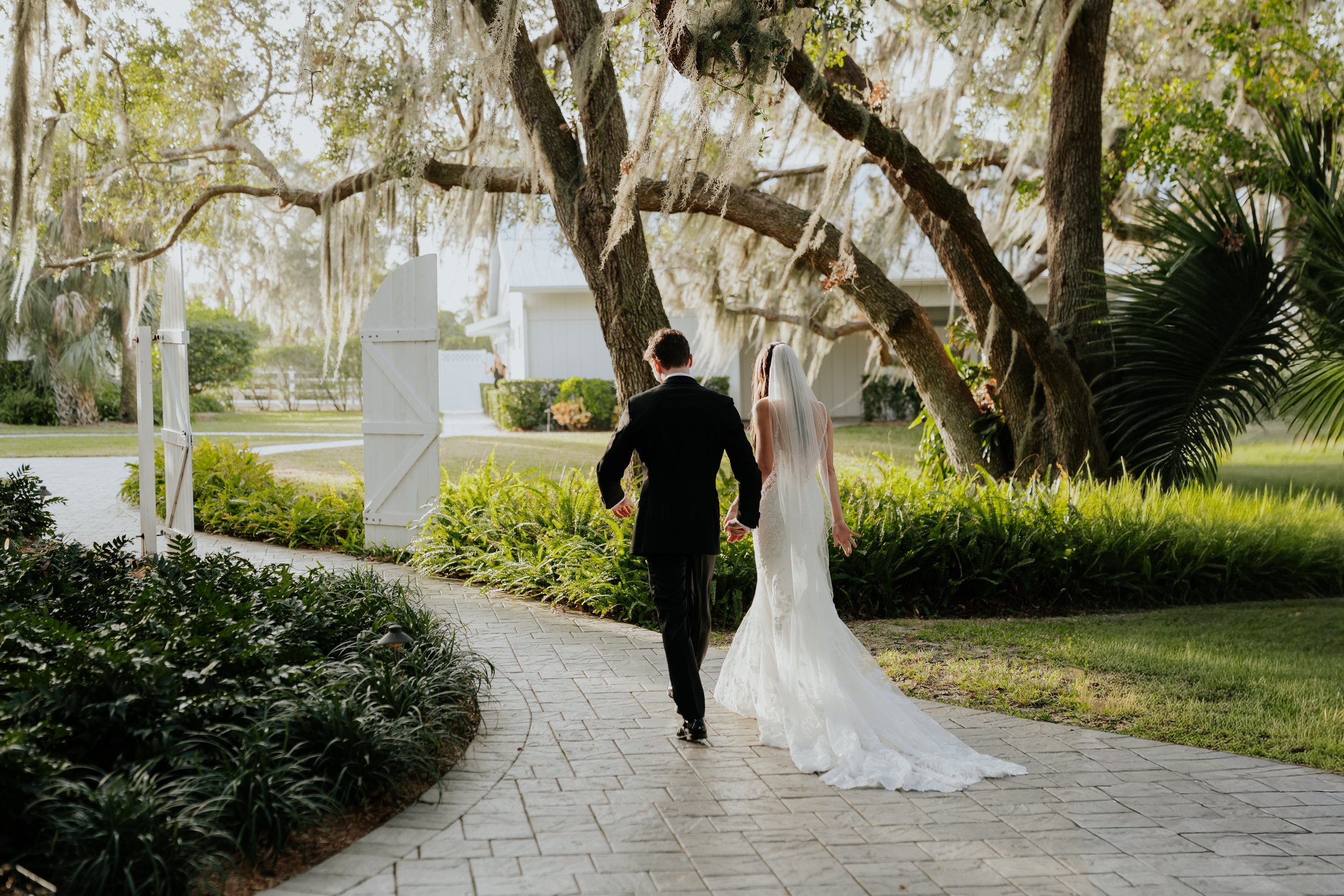A bride and groom holding hands, walking along a stone path under a large tree with Spanish moss, in a garden setting.